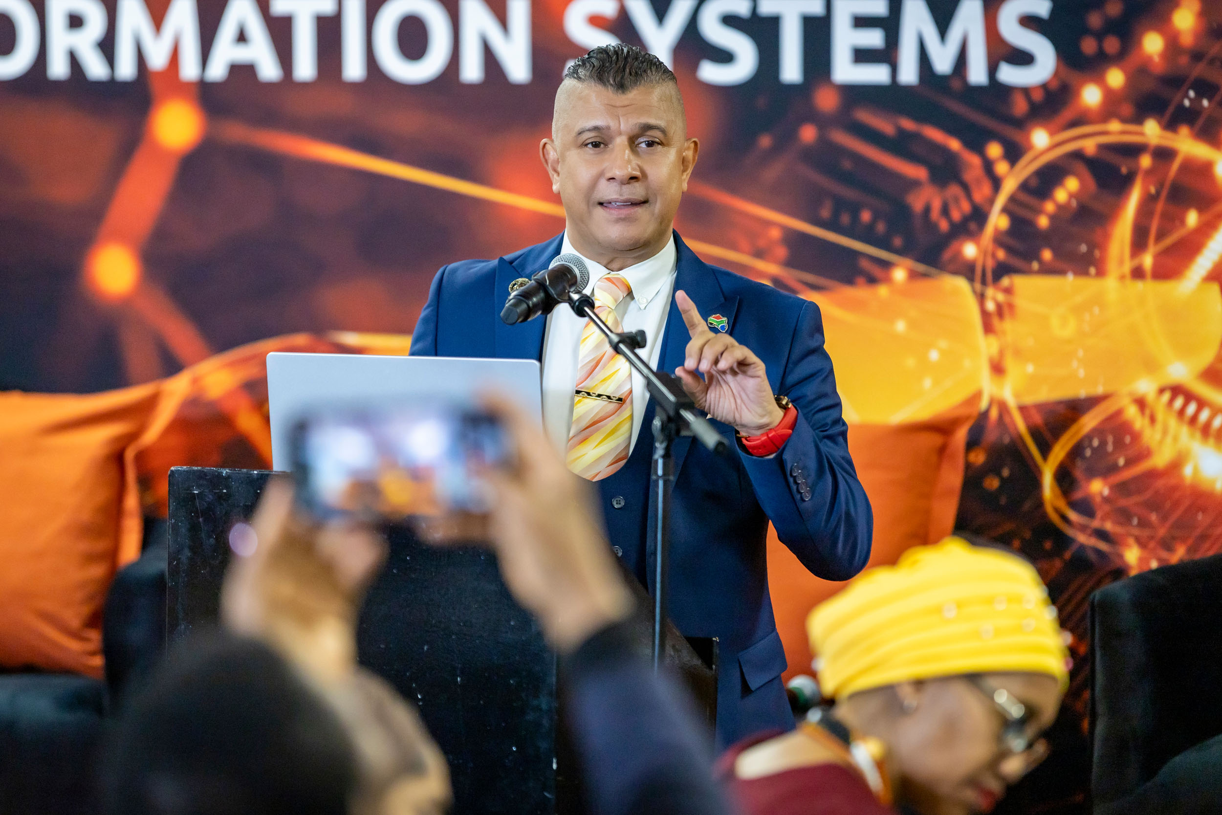 Male speaker in a navy suit and striped tie delivering a talk at a conference, microphones in front, audience blurred in the foreground against an orange tech-themed backdrop.
