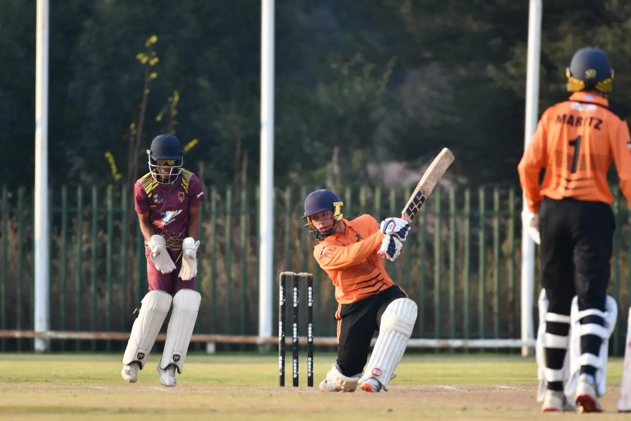 Cricket action: batsman in orange swings at the ball near the wickets as the wicketkeeper in maroon crouches behind the stumps and a fielder watches from the left.