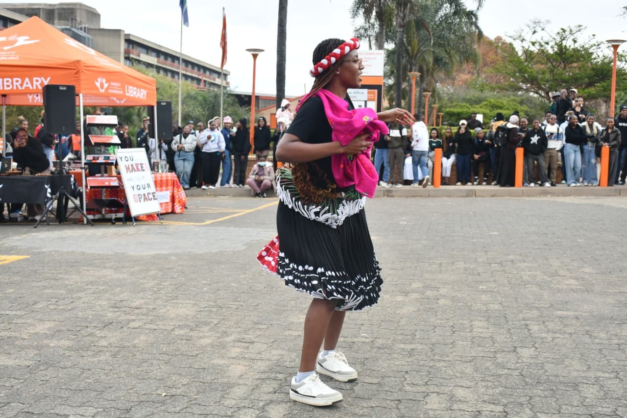 A woman dances in a black and white patterned skirt with a bright pink wrap, wearing a red-and-white headband, at an outdoor community event with a crowd watching.