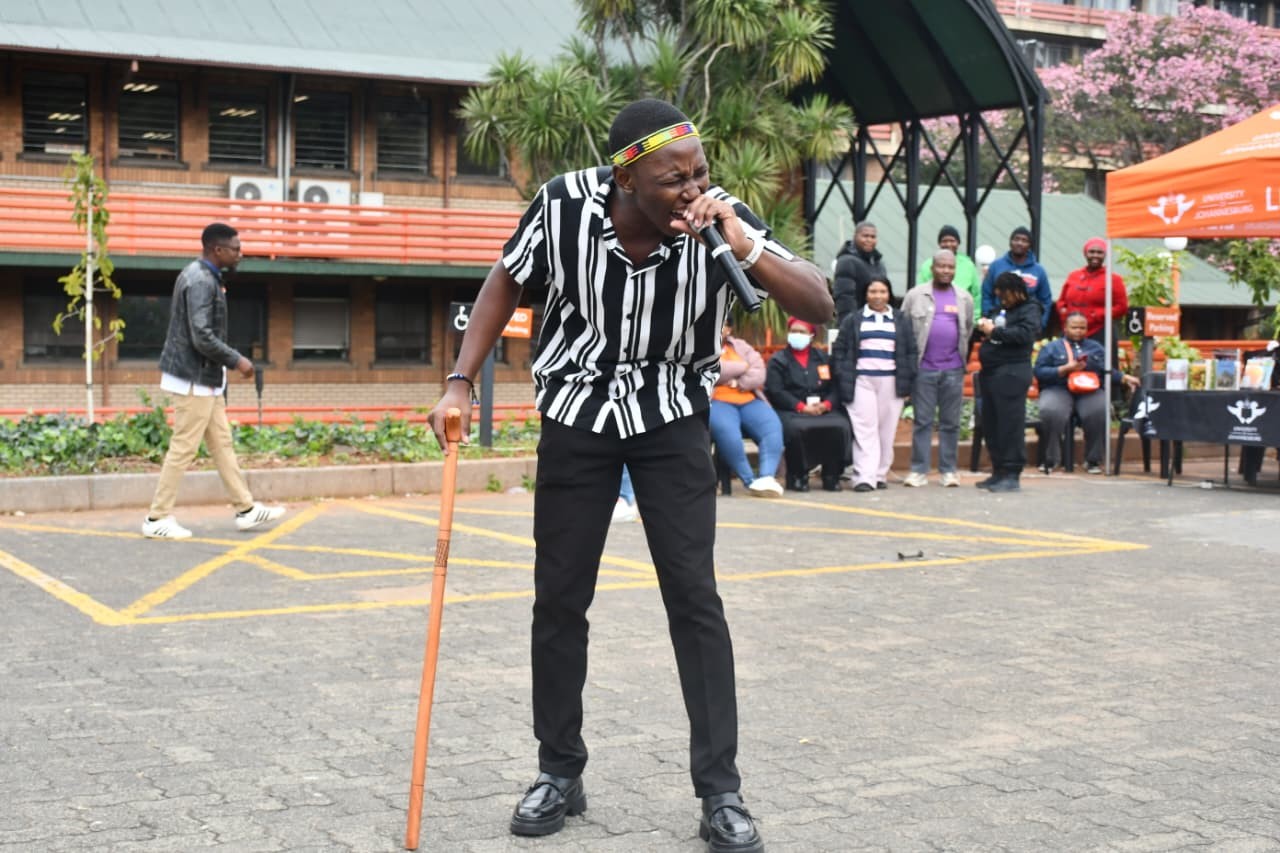 A man in a black-and-white striped shirt and headband sings into a microphone while leaning on a wooden cane at an outdoor event with a crowd watching in the background.