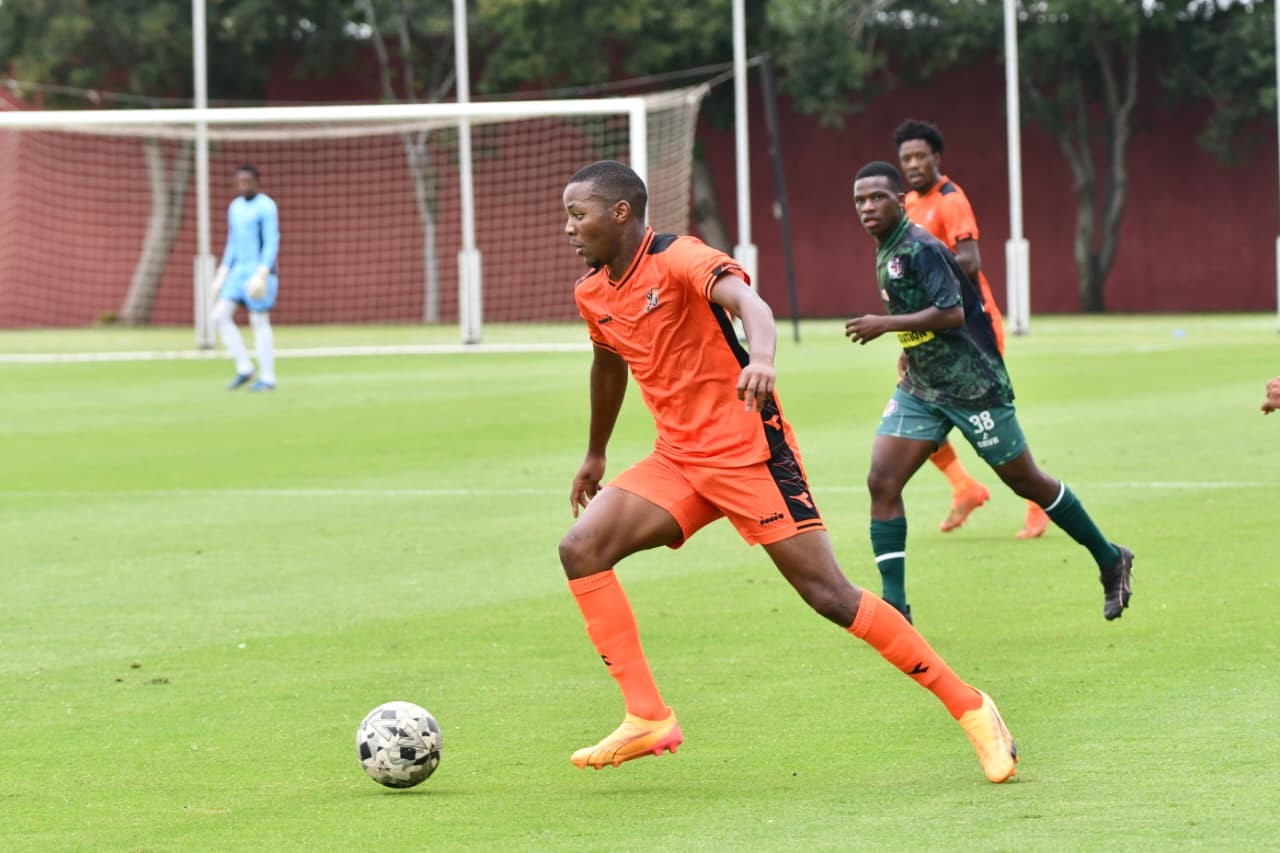 Soccer player in orange controls the ball while others chase on a green field; goalkeeper in blue stands near the goal in the background.