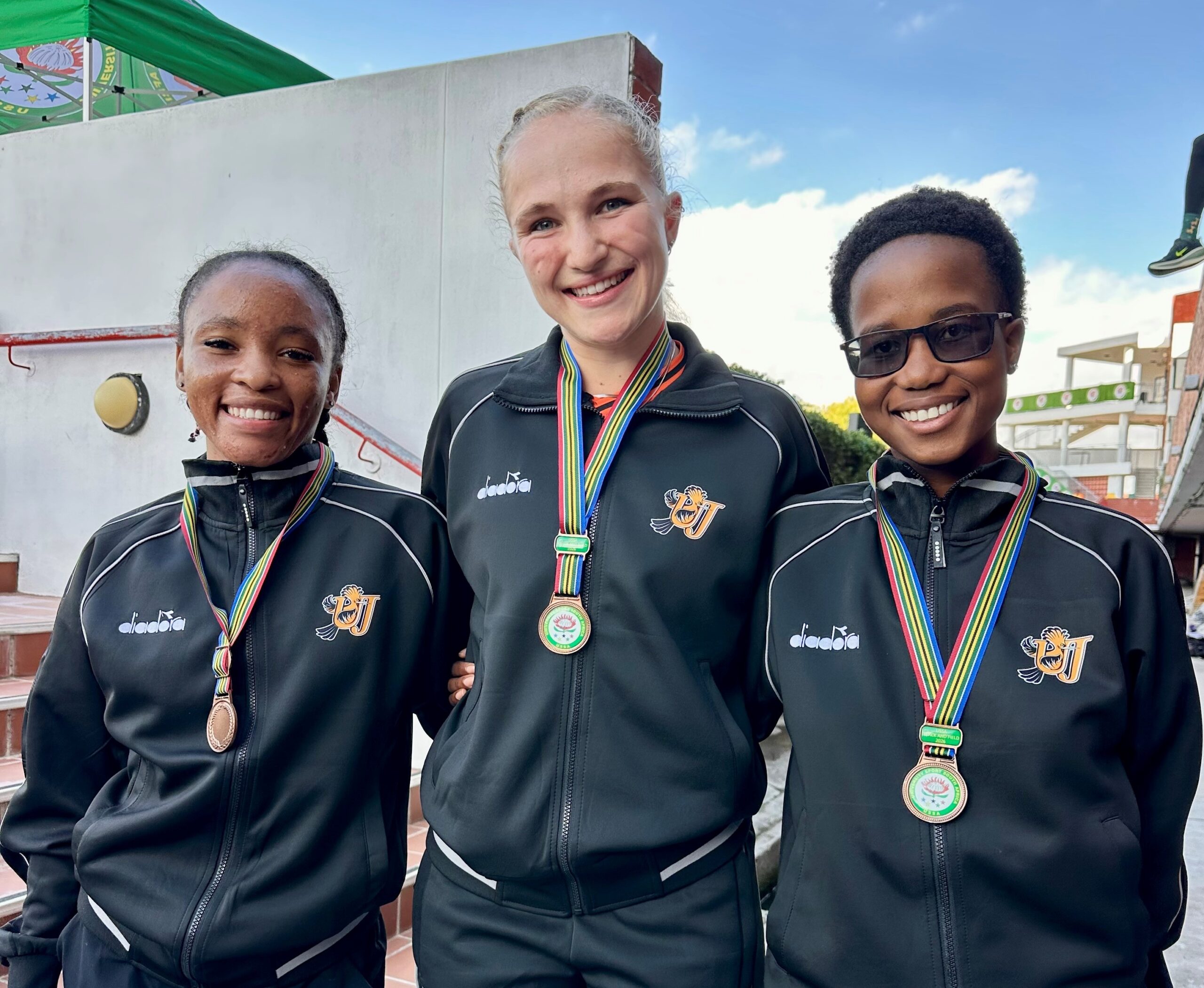 Three smiling athletes wearing matching black jackets with medals around their necks outdoors.