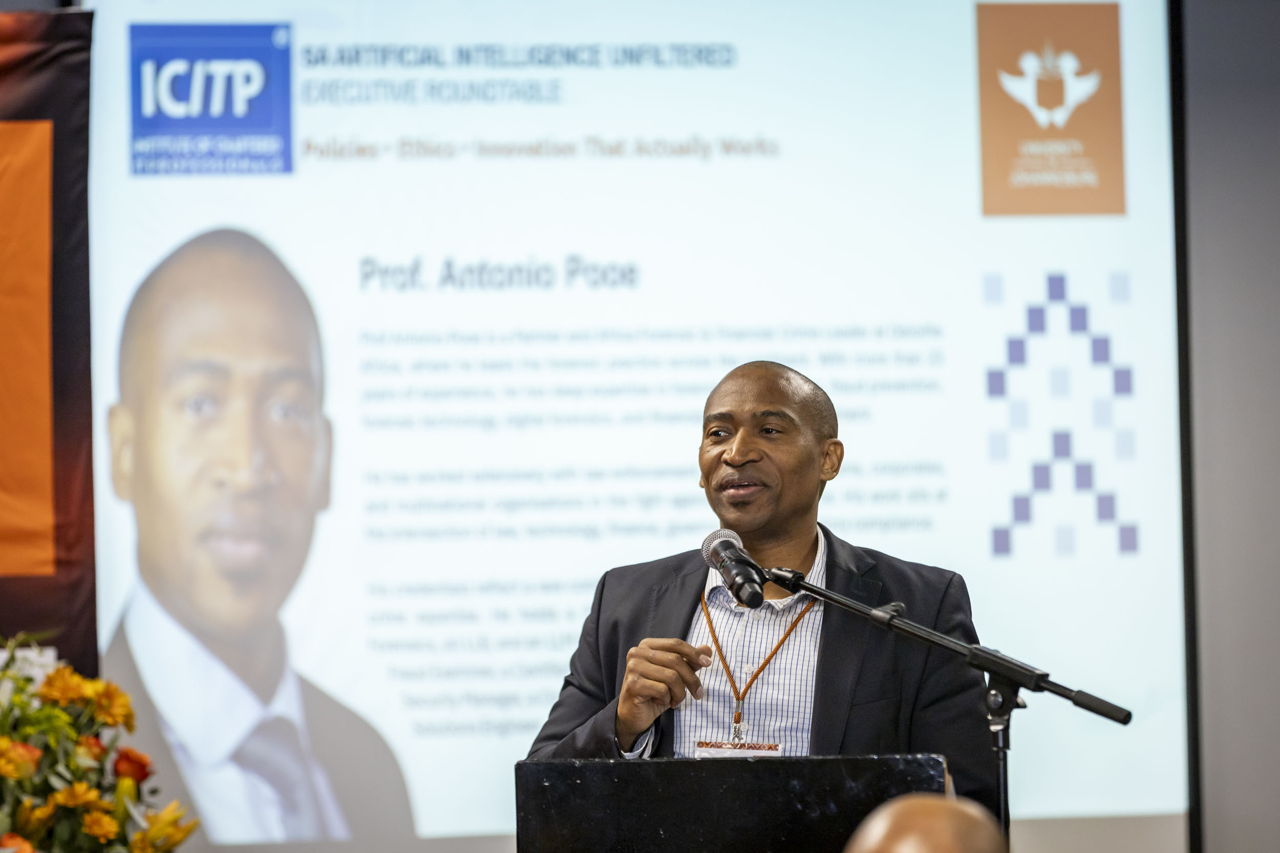 Man in a suit speaks at a podium with a microphone, standing before a large presentation screen with an ICTP logo and a portrait on the screen behind him.