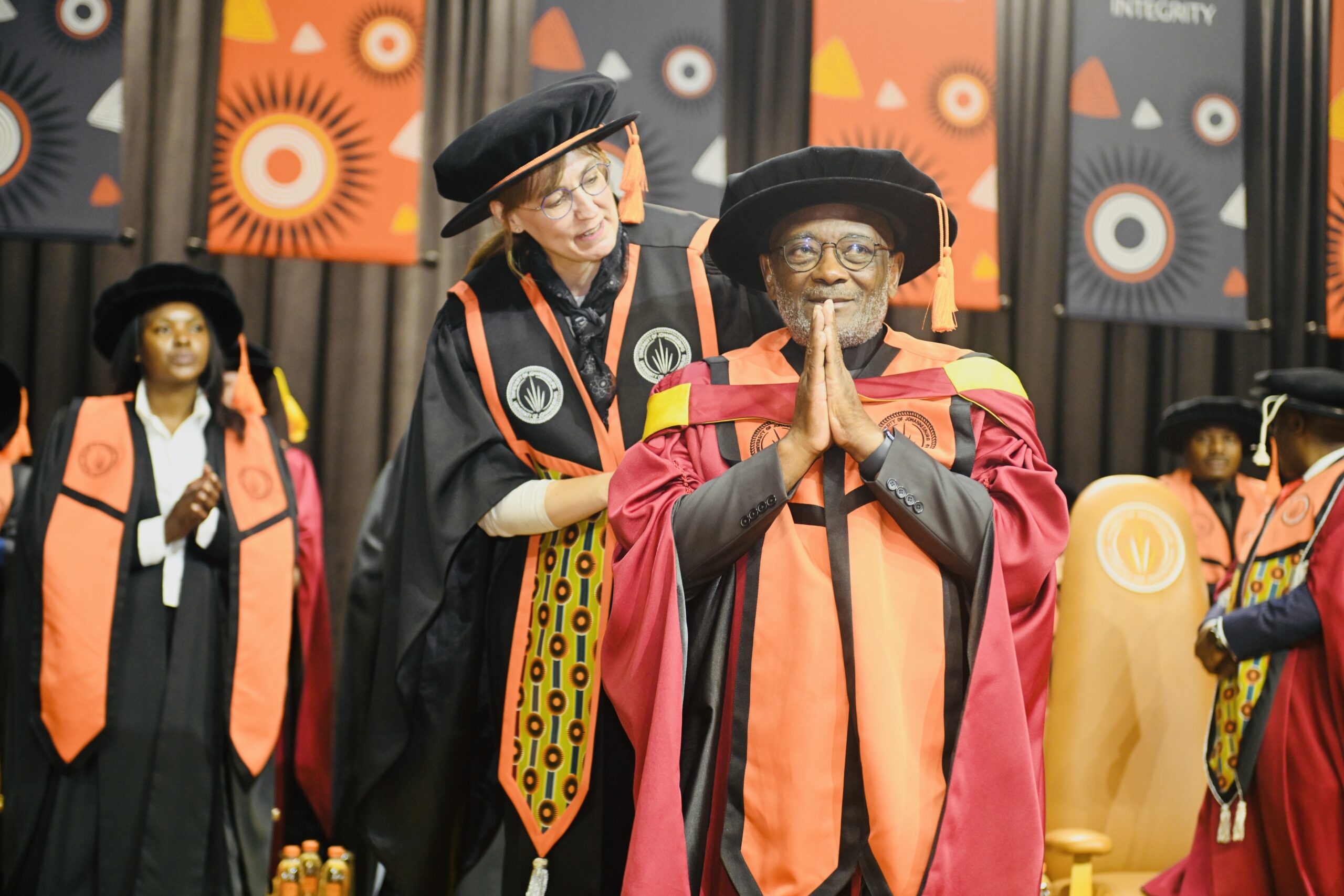 Elderly graduate in pink-red gown and black cap with hands pressed in prayer, smiling on stage amid bright banners and fellow grads.