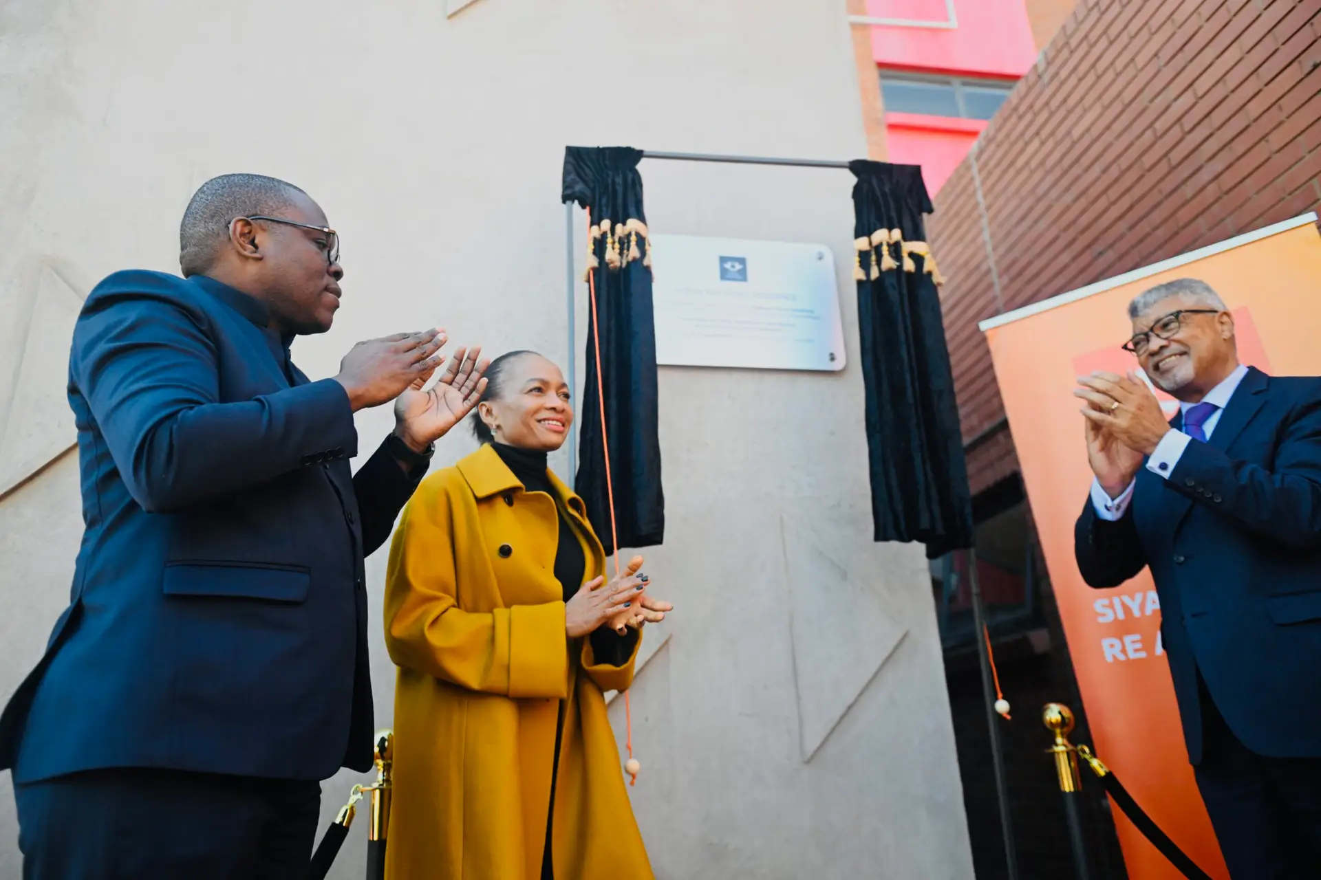 People at an unveiling ceremony clapping beside a plaque on a beige wall, with black curtains and an orange-banner backdrop behind them.