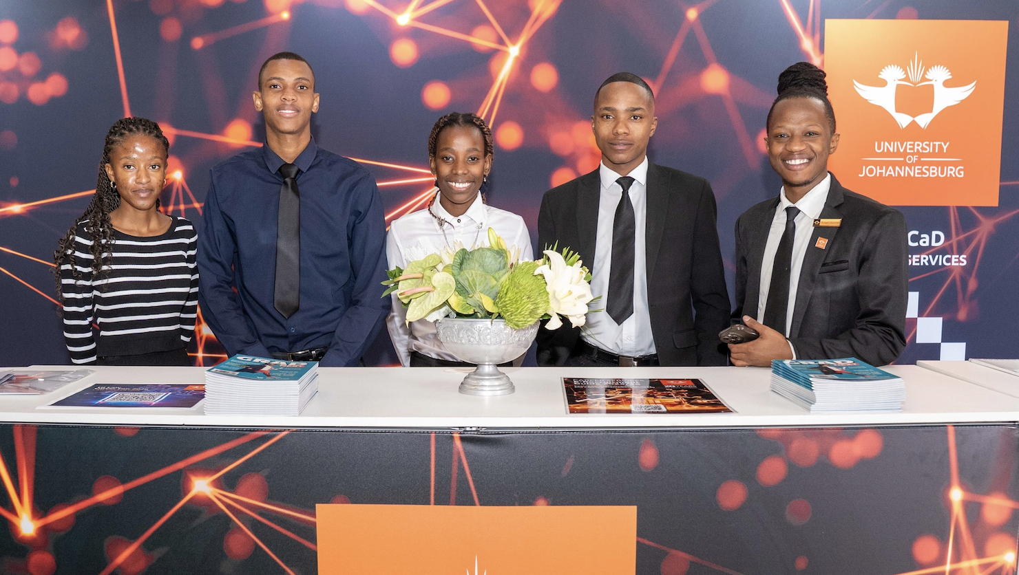 Five students in business attire stand behind a welcome desk at the University of Johannesburg, with brochures and a floral centerpiece.