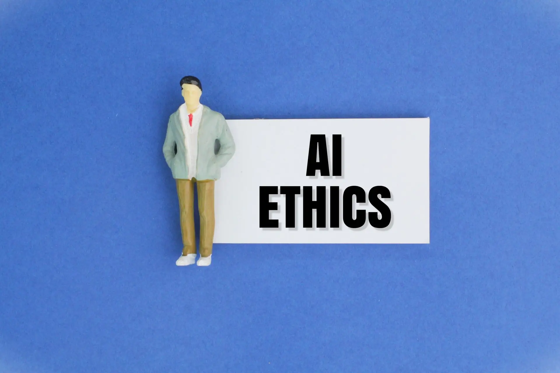 Small figurine of a man in a pale jacket standing beside a white card that reads 'AI ETHICS' on a blue background.