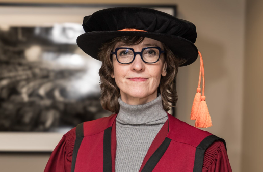 Professor in burgundy academic gown and black hat with orange tassel smiles at the camera.