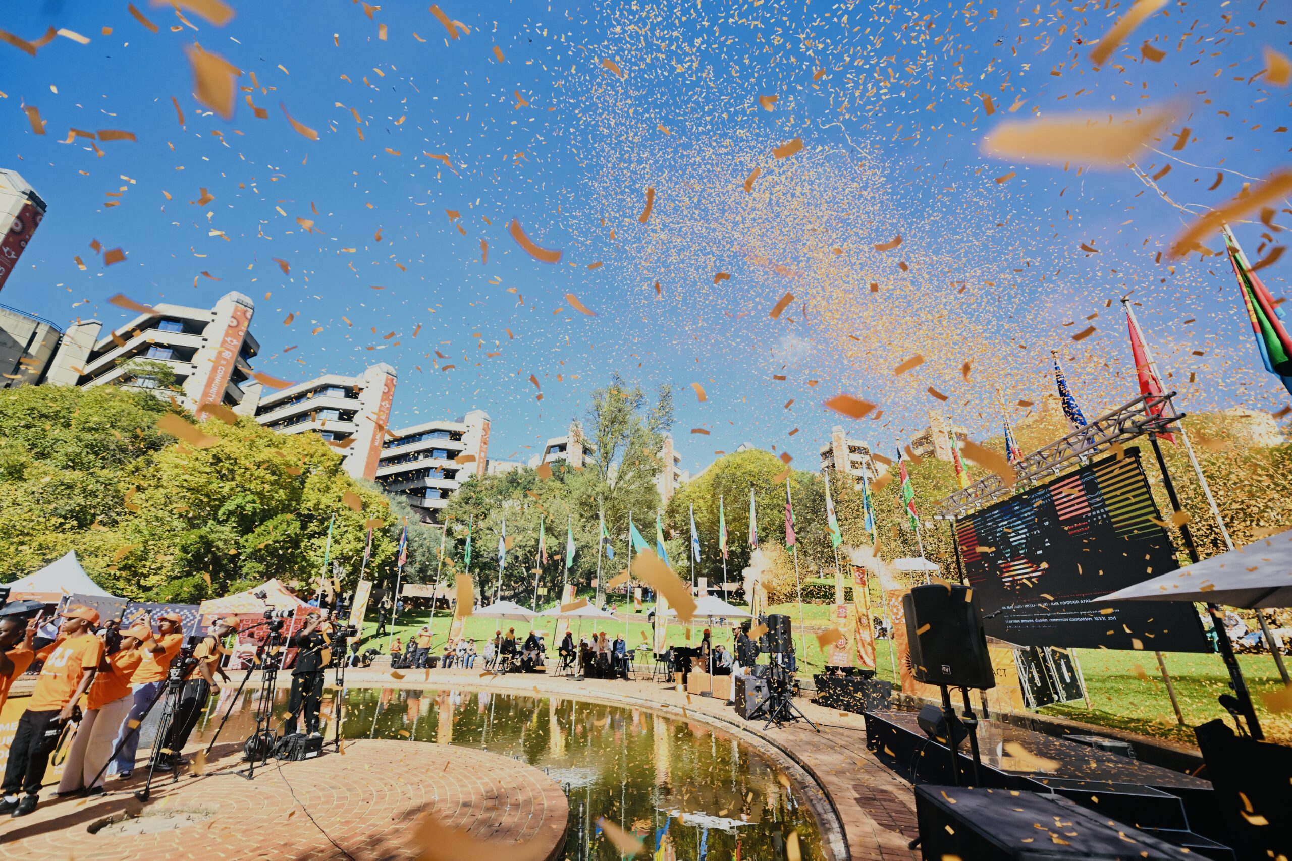 Outdoor celebration with orange confetti raining over a stage and audience beneath a blue sky, with colorful flags and buildings in the background.