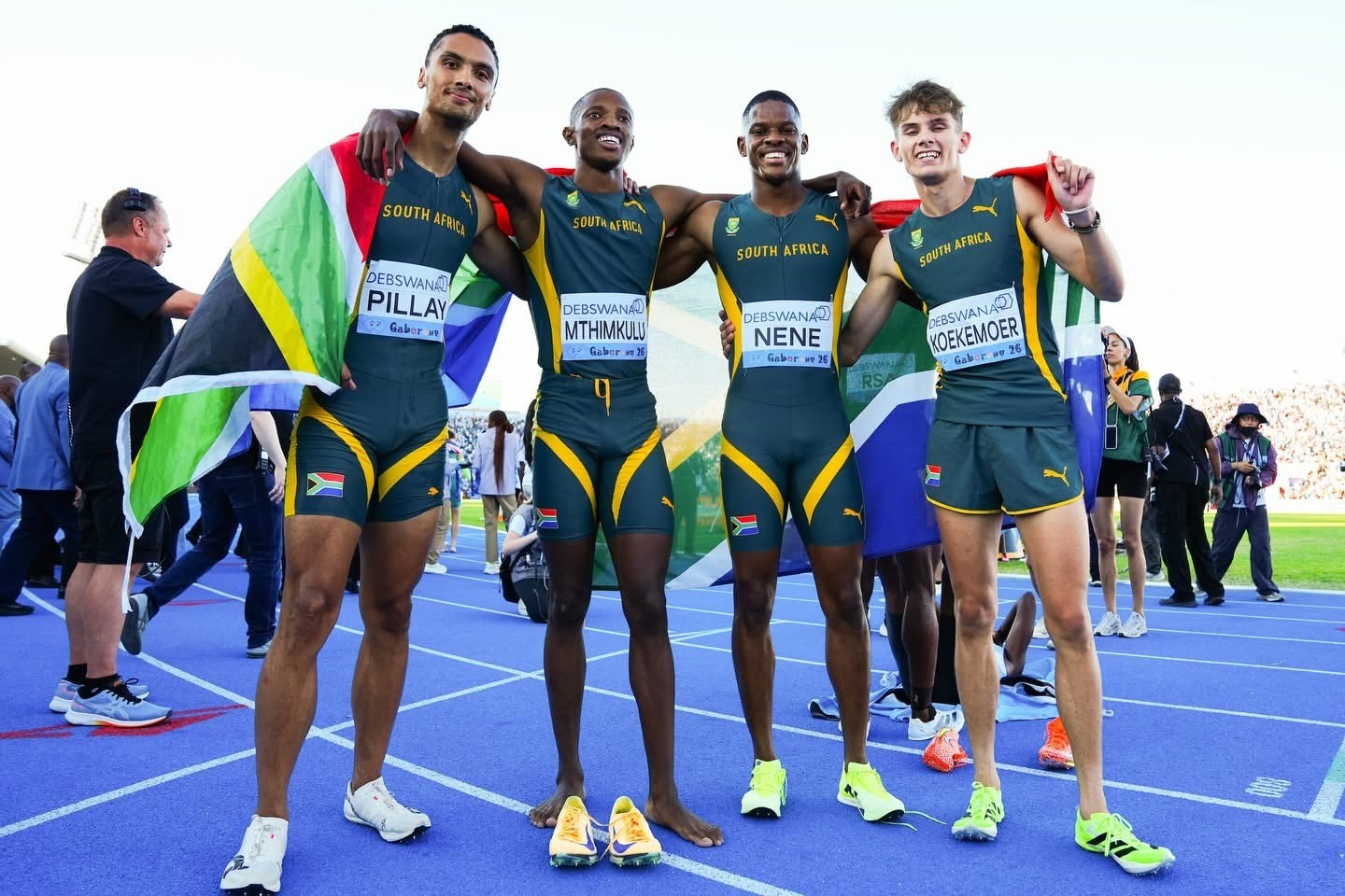 Four South African male sprinters stand arm-in-arm on a blue running track, draped in national flags and smiling for the camera.