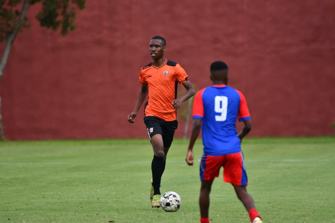 Soccer players on a green field; an orange-jersey player dribbles the ball while a blue-and-red defender approaches.