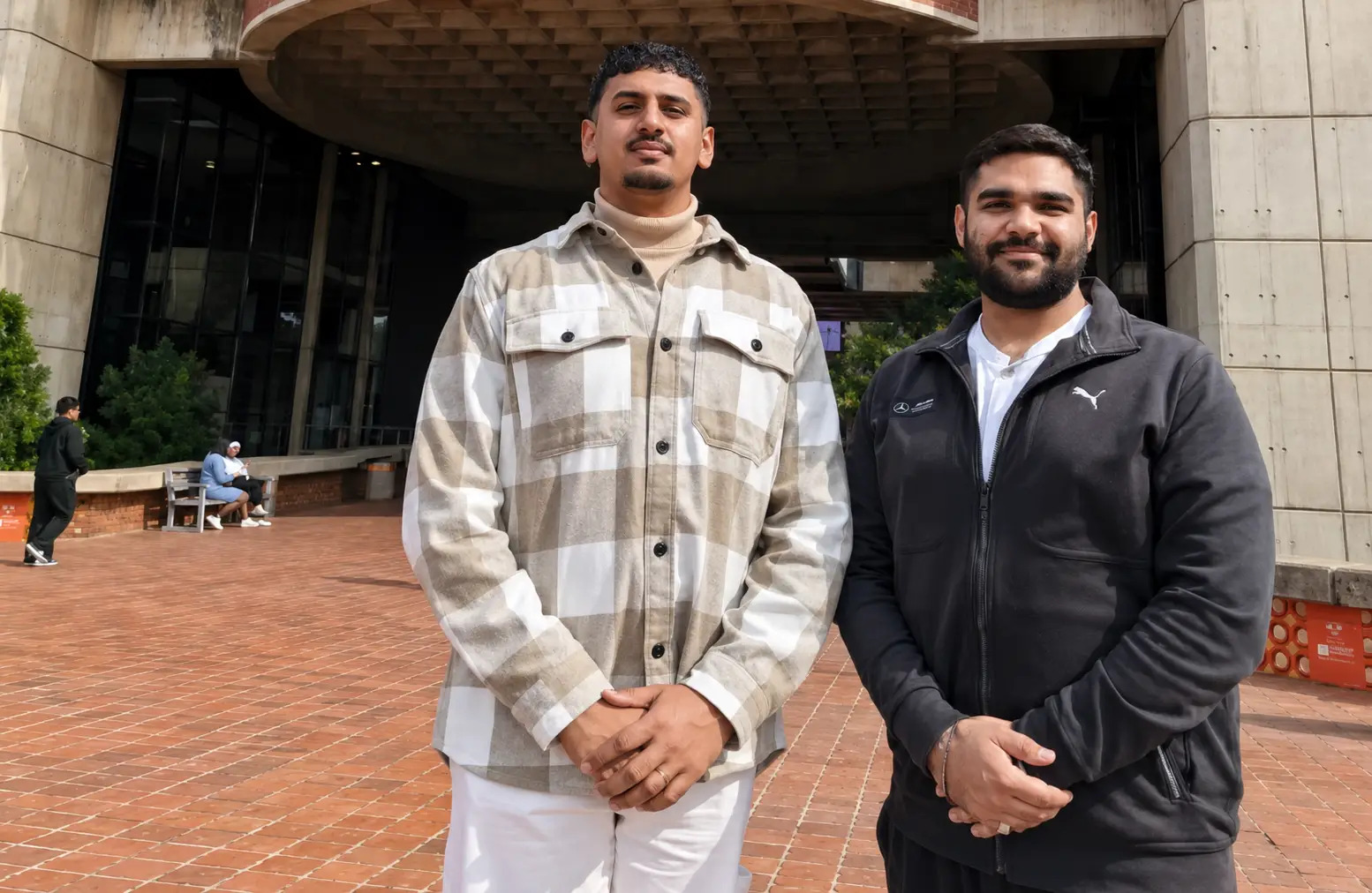 Two men stand outdoors in front of a modern building with a brick-paved plaza: one in a beige plaid shirt, the other in a black zip-up jacket, both smiling.