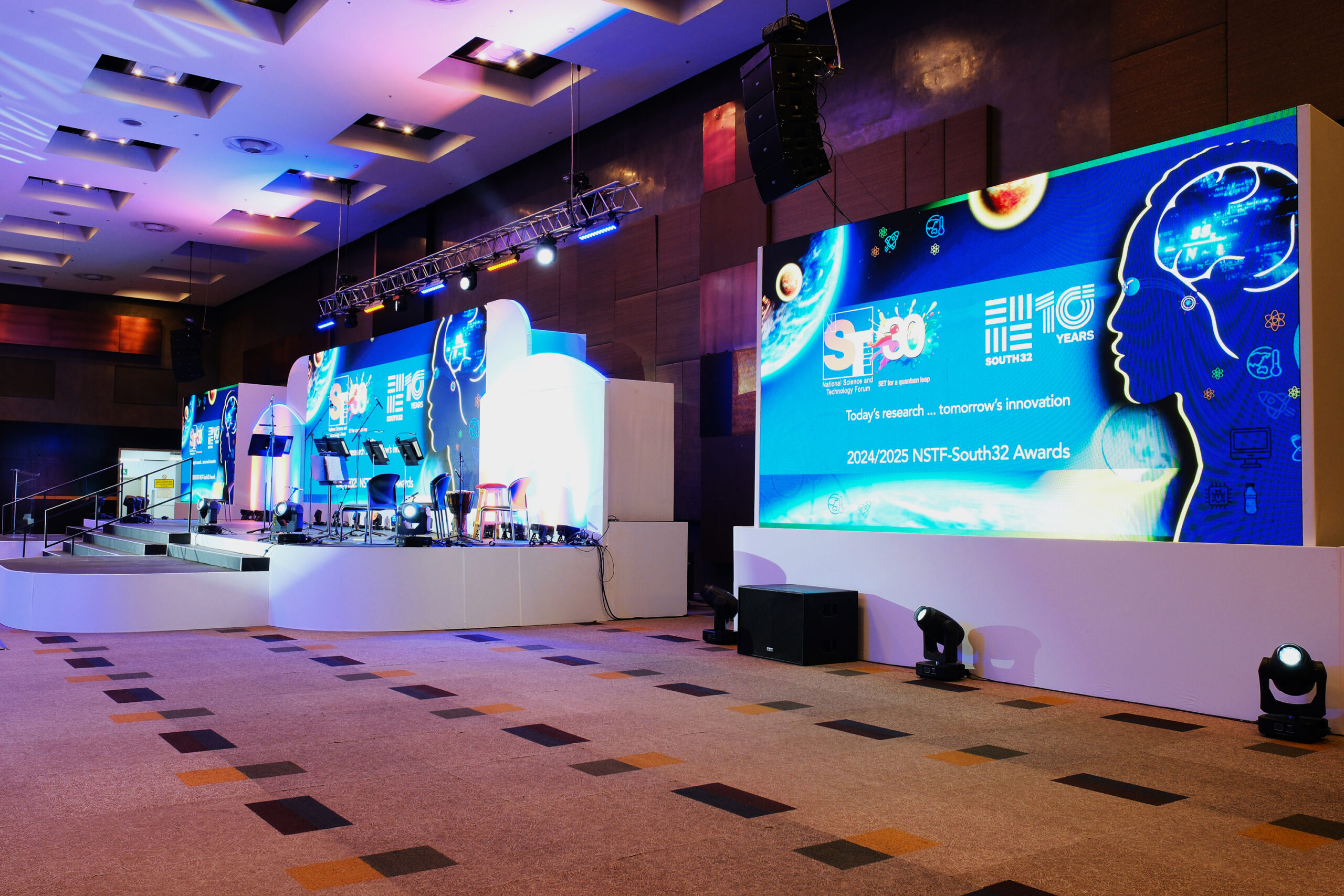 Stage setup for a science/tech conference with a large blue digital backdrop, seating, and lighting rig on a multi-level platform.