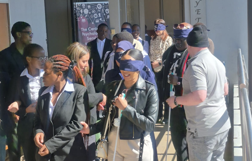 Diverse group of blindfolded participants lined up outside a building, some guiding each other during an activity.