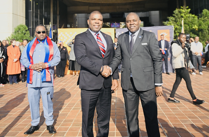 Two men in dark suits shake hands in a brick courtyard outside a modern building, with onlookers in the background.