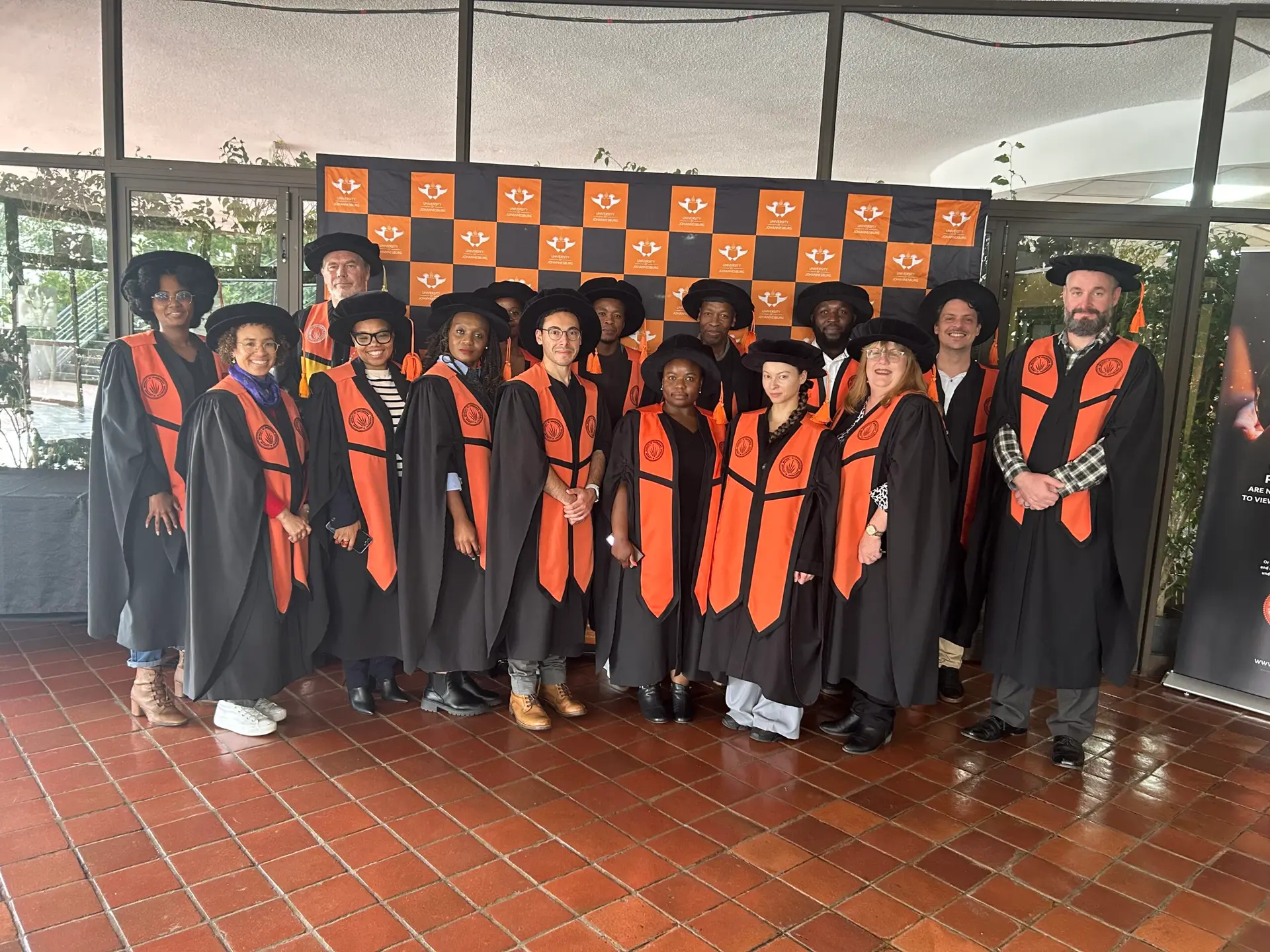Group of graduates in black gowns with orange sashes posing indoors in front of an orange-blue branded backdrop.