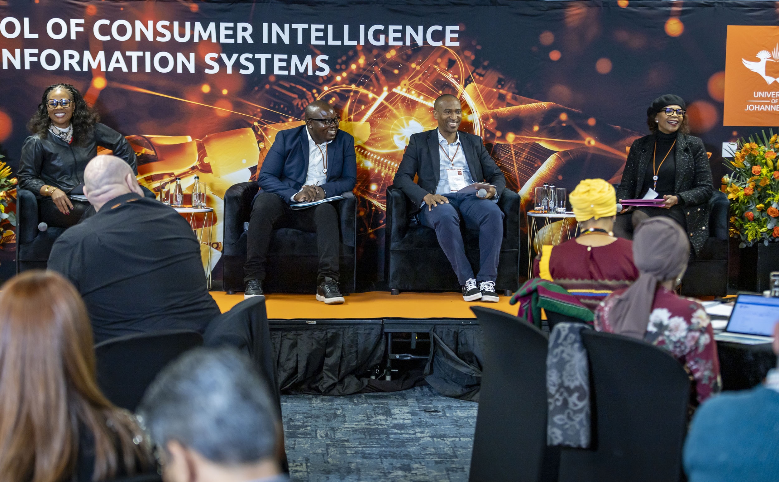 Panel discussion with four speakers seated on a stage, smiling, presenting to an audience in a conference hall; vibrant orange and tech-themed backdrop behind them.