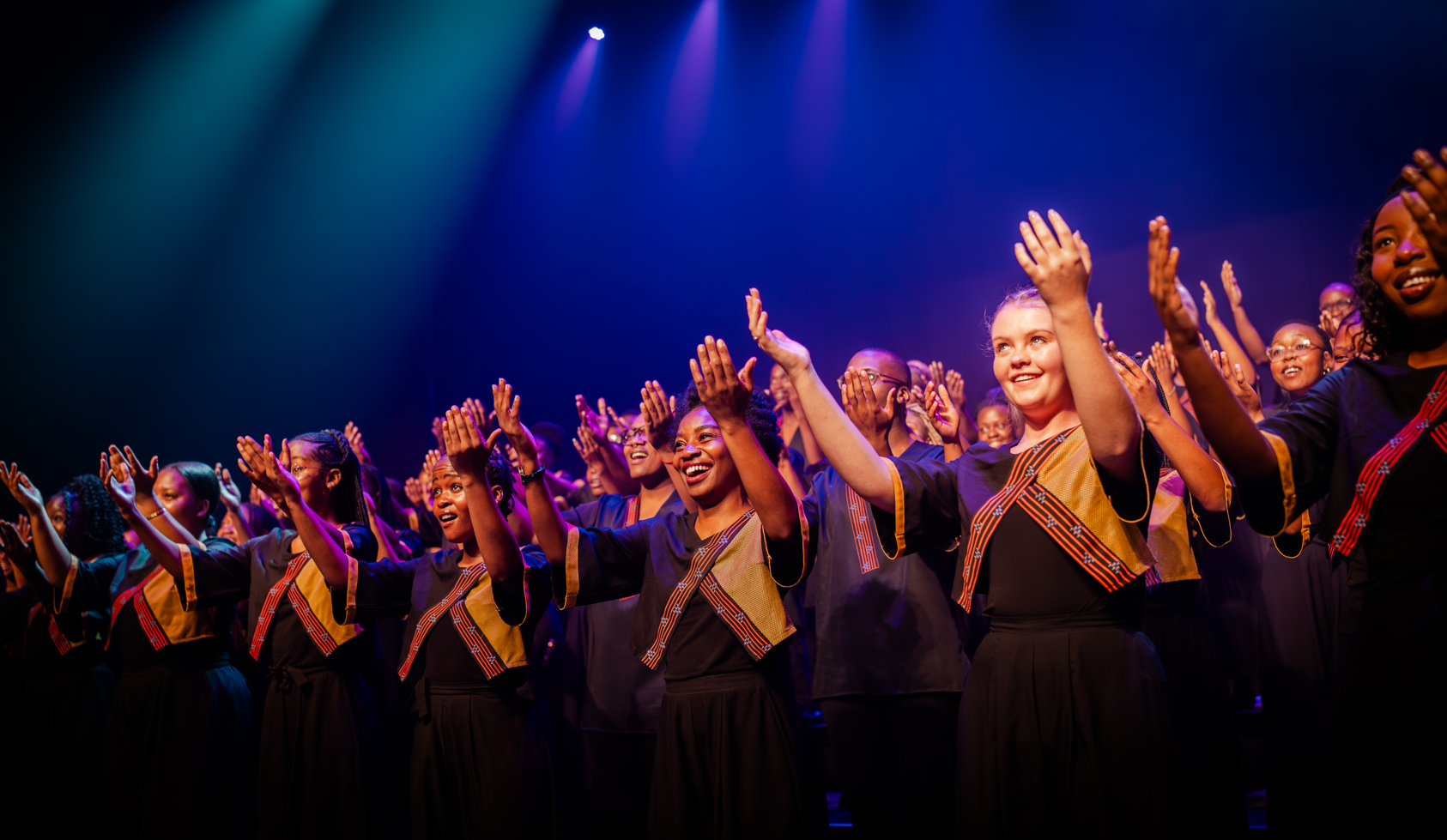 A large choir on stage with colorful sashes, raising their hands in unison during a performance under blue and purple lights