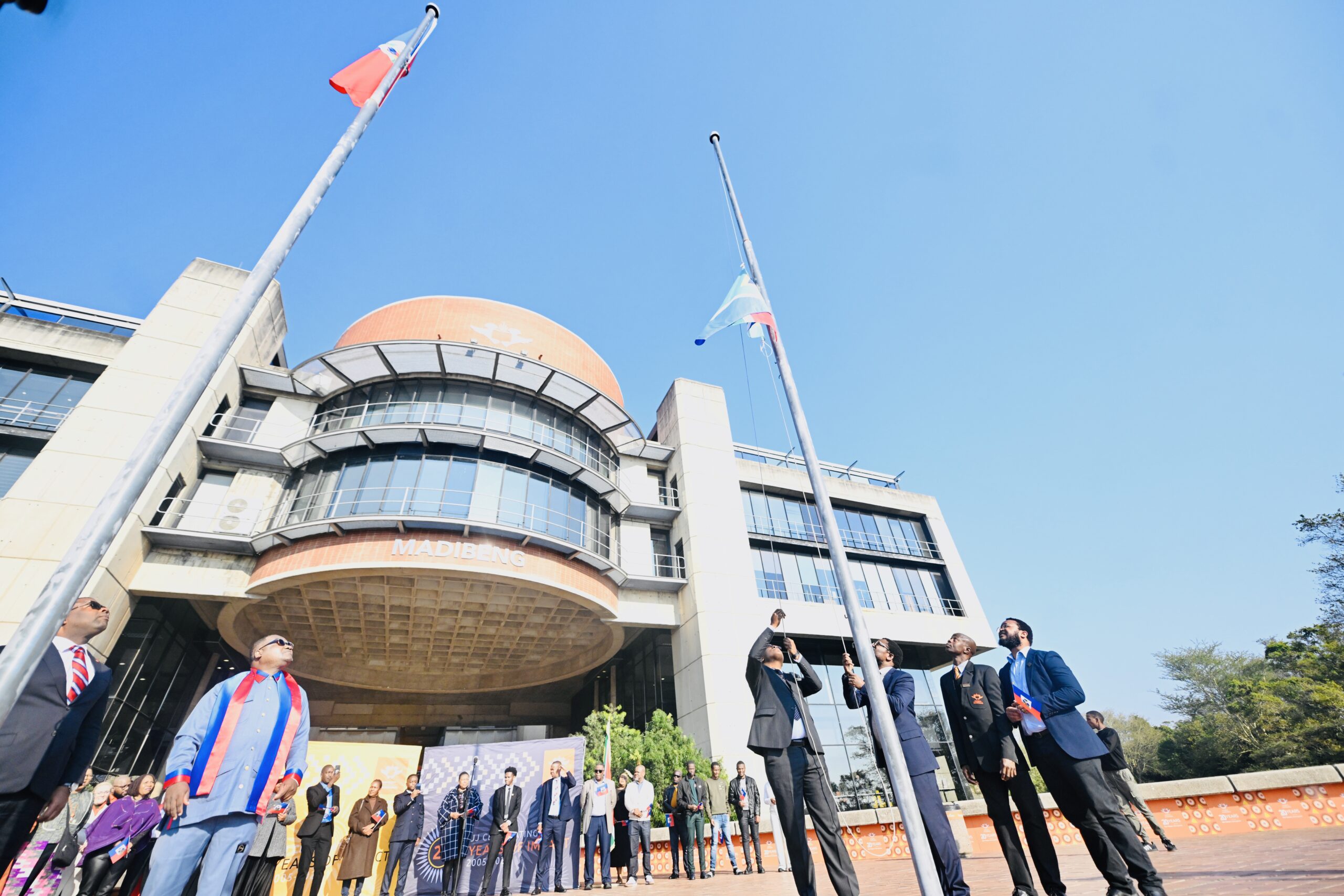 Group of professionals in suits standing outside a modern building with flagpoles and an orange-brick facade near the entrance.