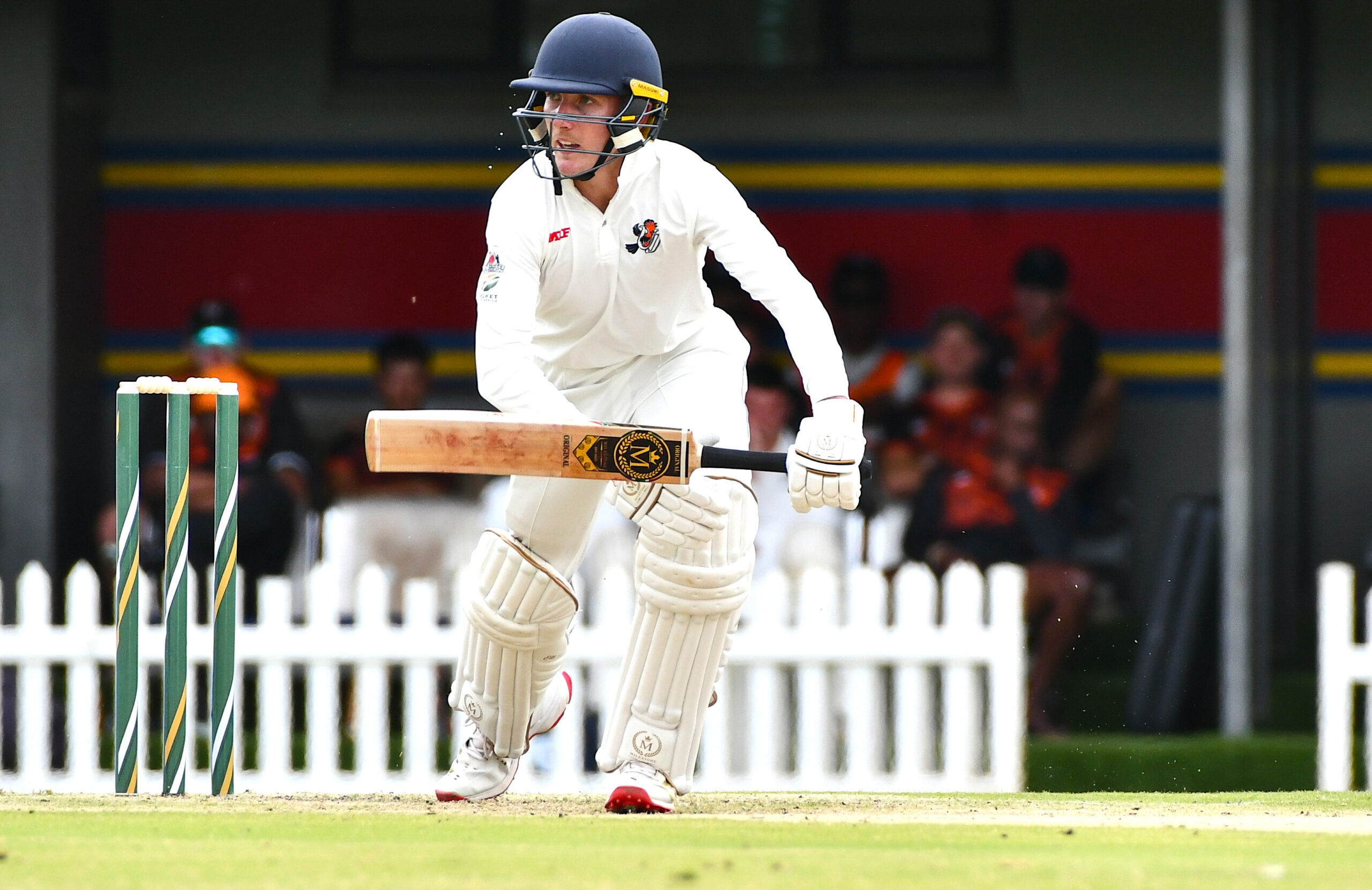 Cricket batsman in white gear drives the ball, helmet on, near the stumps on the left.