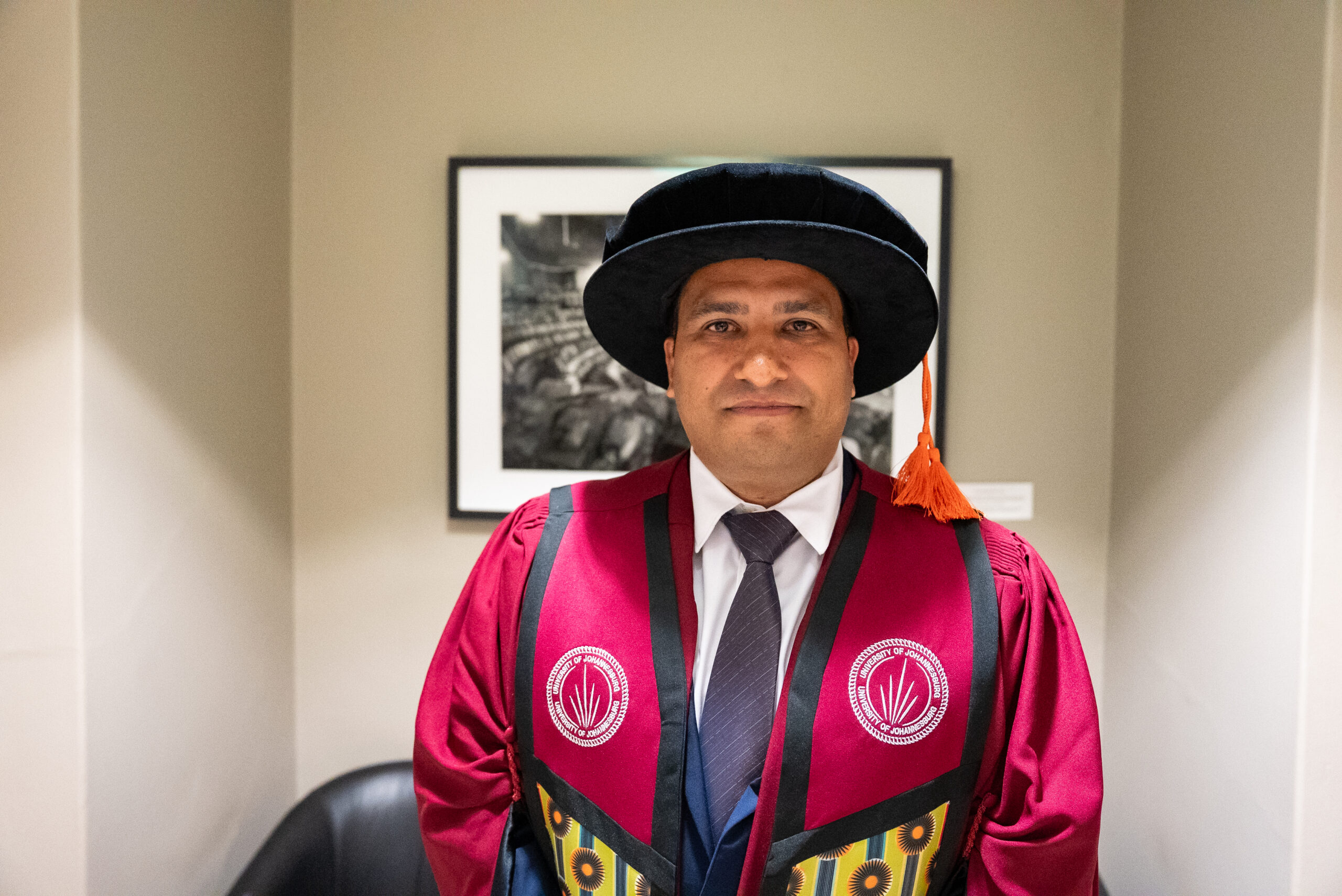 Graduate in a maroon gown and black academic cap poses in a hallway with a framed photo behind him.”] ,