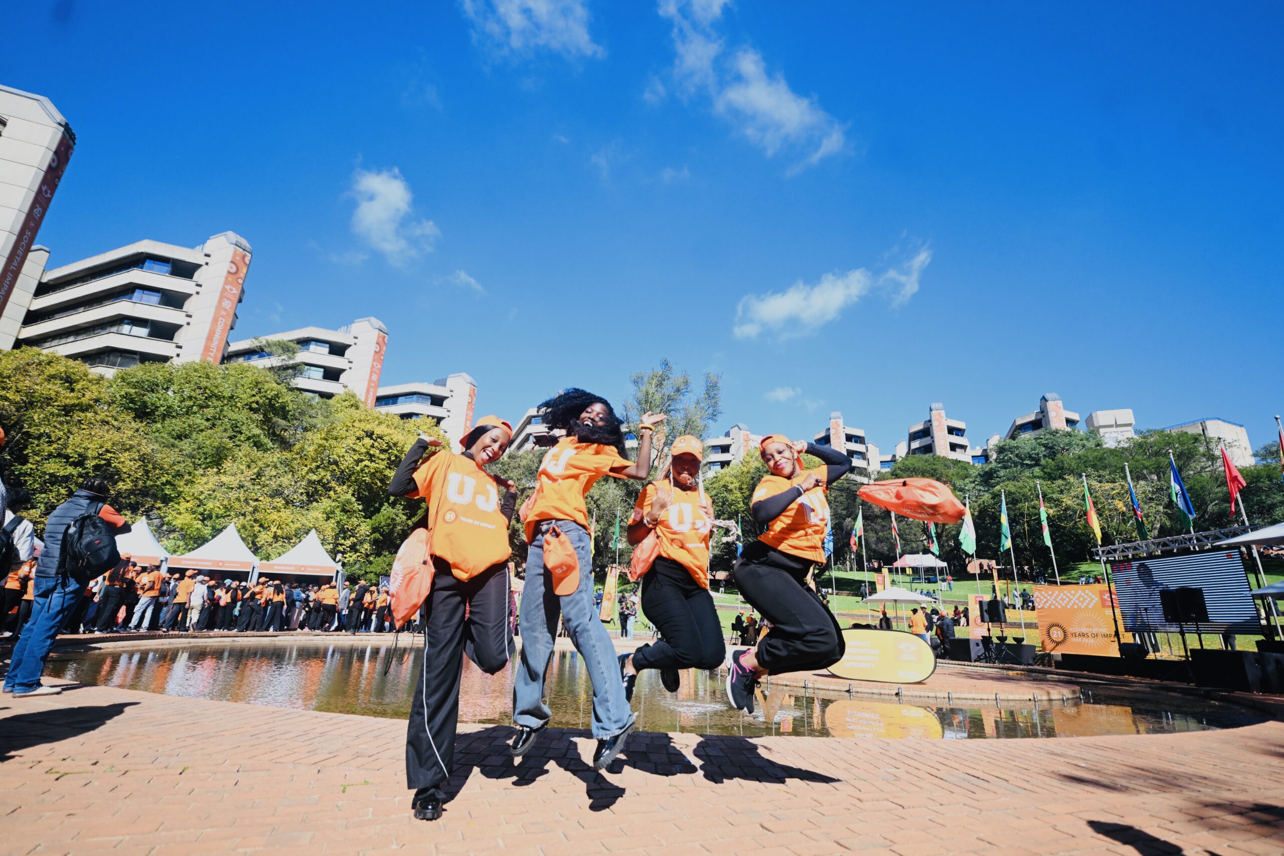 Group of people in orange shirts jumping in a plaza during an outdoor campus event, with blue sky and buildings behind.