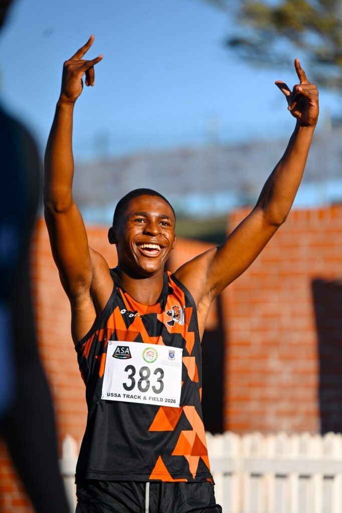 Young male runner on a finish line raising both arms in celebration, wearing a black and orange geometric singlet with bib 383