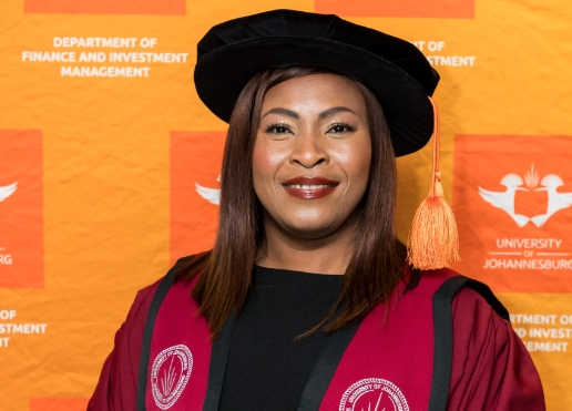 Smiling woman in a maroon doctoral gown and black academic cap at a university ceremony, with an orange backdrop.