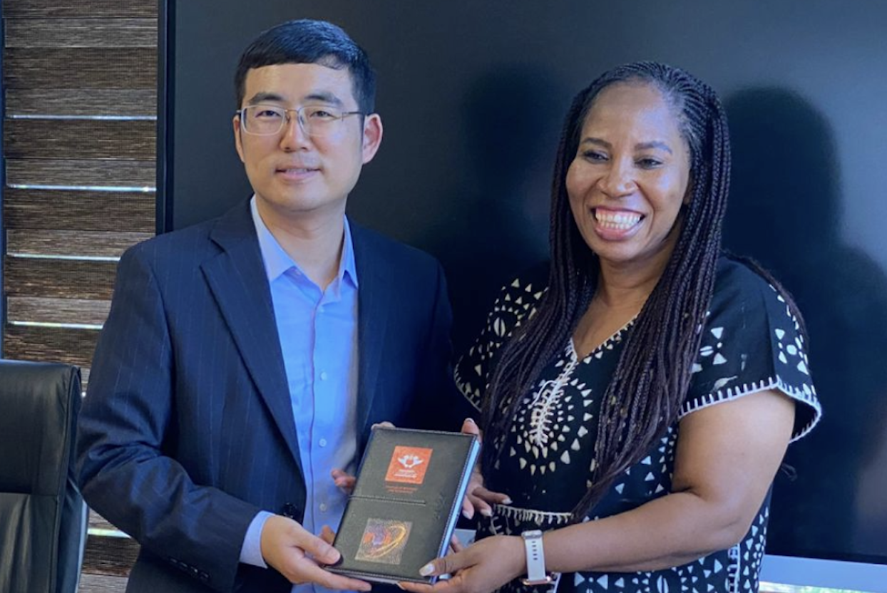 Man in a blue suit presents an award plaque to a smiling woman in a patterned dress inside an office.