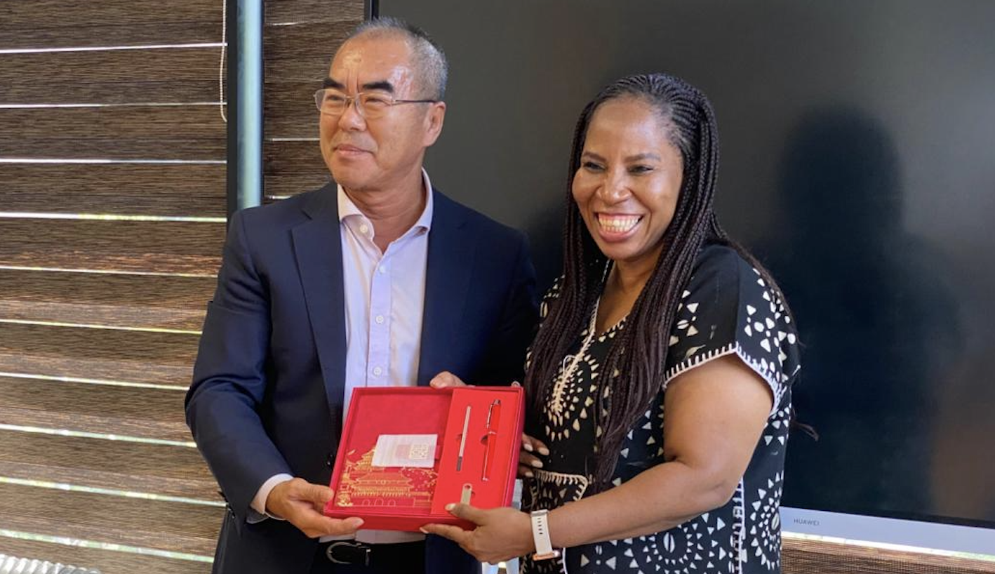Man in a dark suit and a smiling woman exchange a red gift box containing pens for a formal presentation.