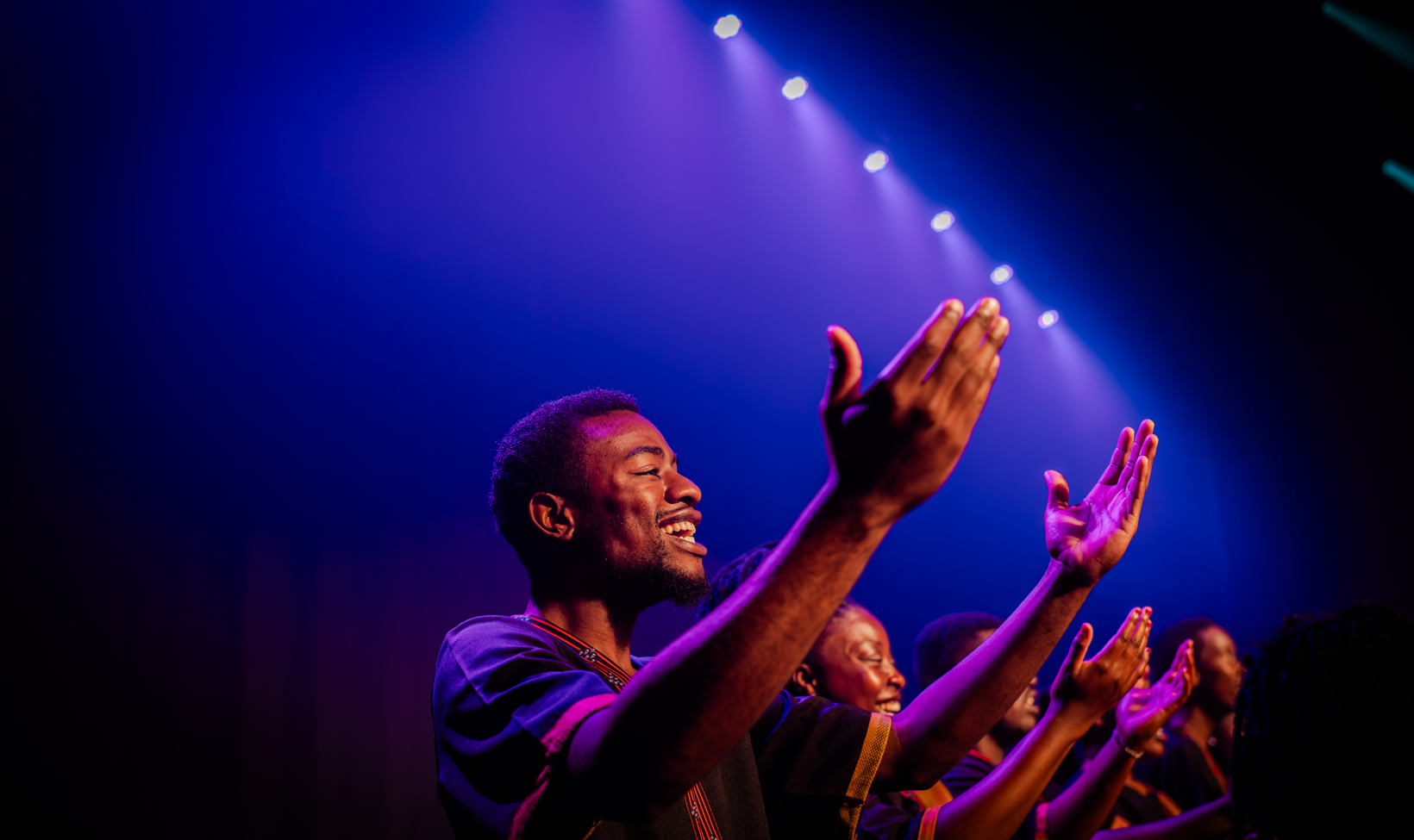 Group of people on a stage clapping and smiling under purple and blue concert lights accessibly described as a live performance moment.
