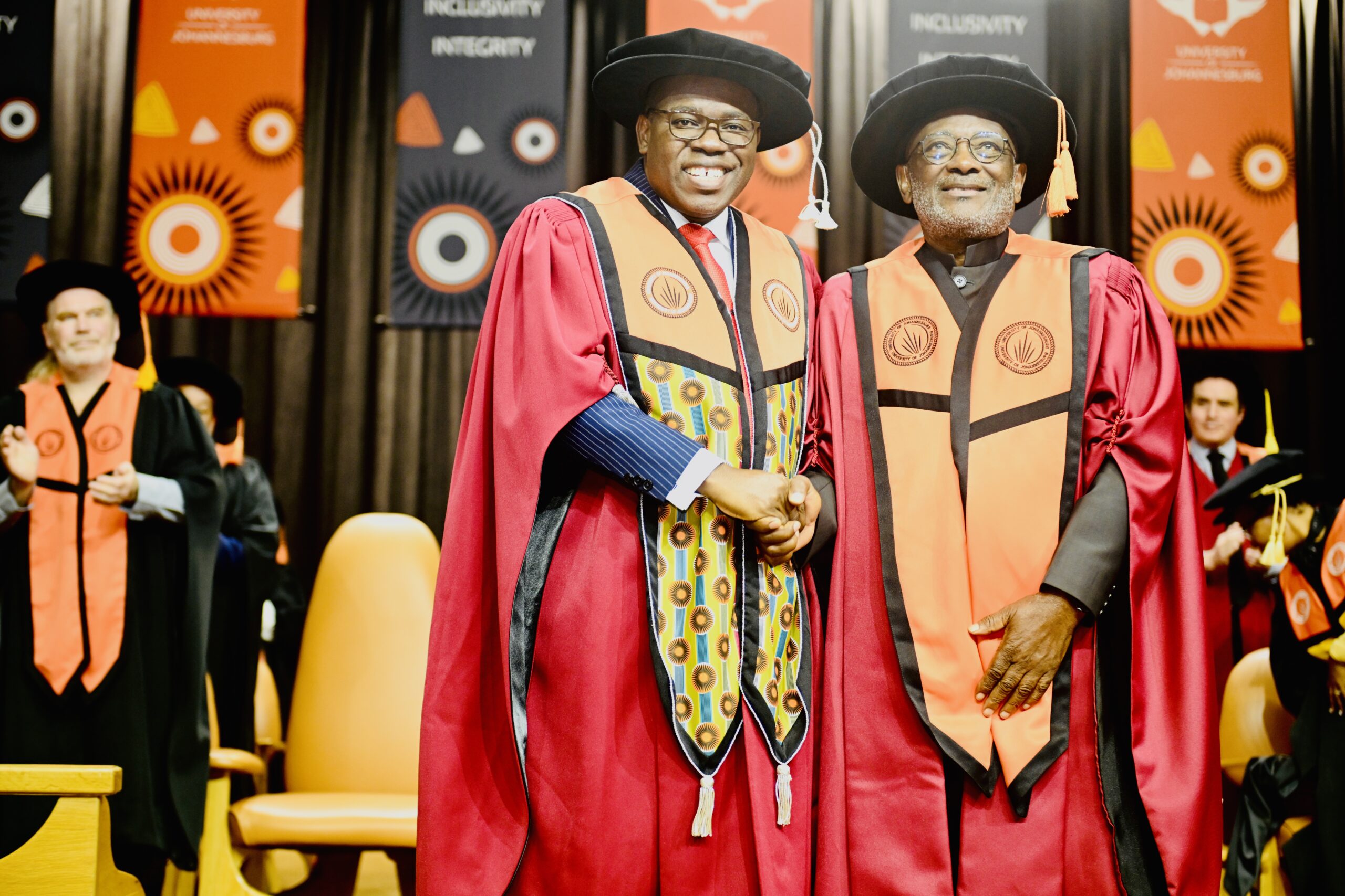 Two graduates in red gowns and black hats shake hands on a stage during a ceremony, with orange and dark banners featuring circular designs in the background.