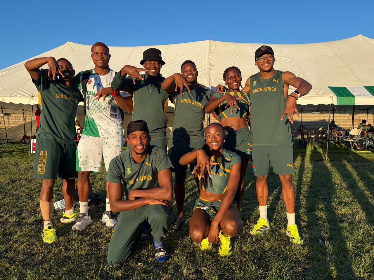 South African track team posing for a photo outdoors in green uniforms on a sunny grassy field with a large white tent in the background.