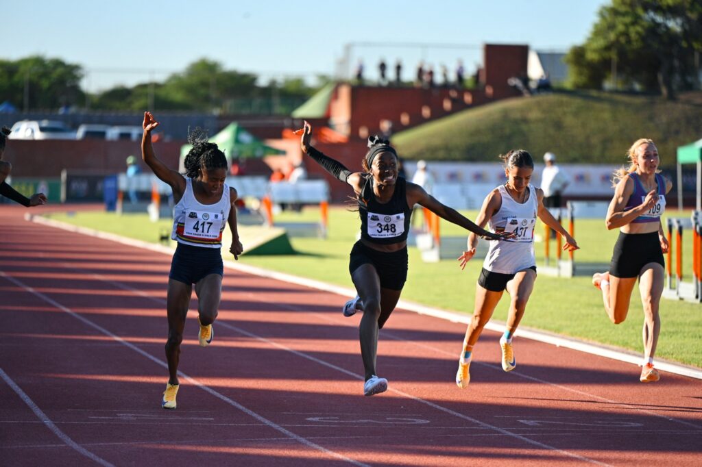 Several female sprinters race down a red track, arms pumping and bib numbers visible (417, 348, 431).
