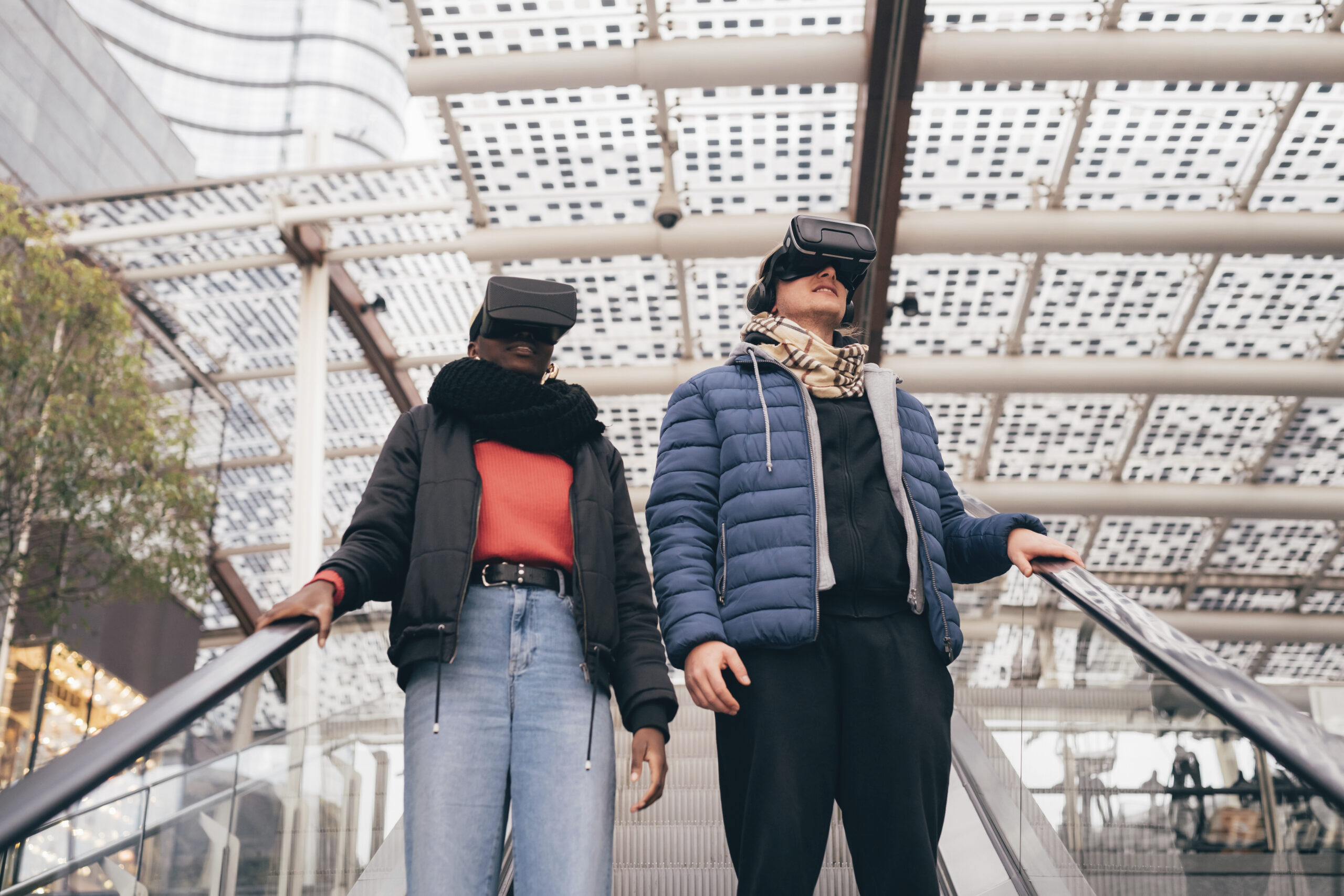 Two people on an escalator wearing VR headsets in a glass-walled mall.