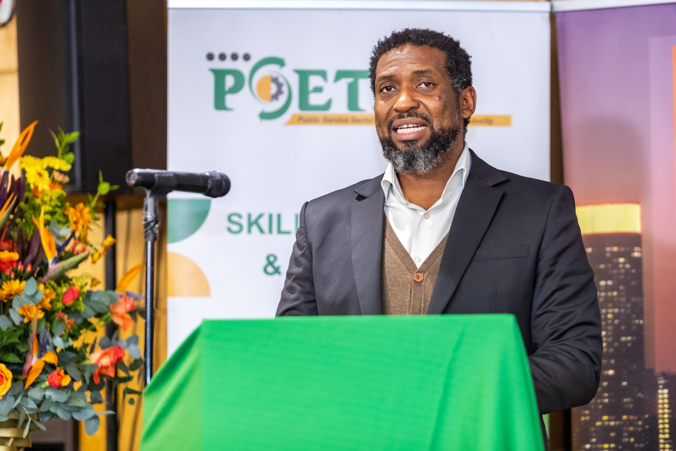Male speaker in a suit delivering a talk behind a bright green podium, with a banner and colorful flower arrangement in the background.