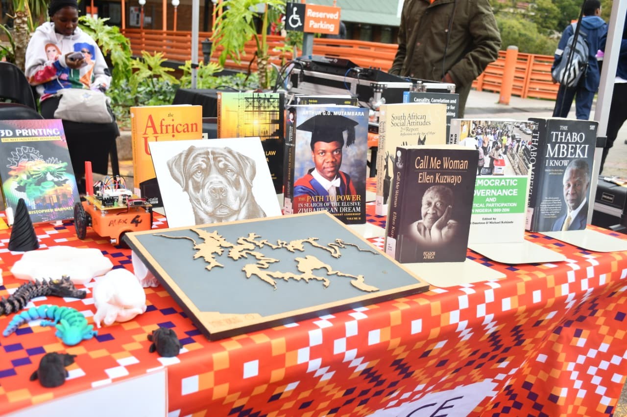 Outdoor table of books and 3D-printed objects on a red-orange checkered cloth, with a Reserved Parking sign in the background.