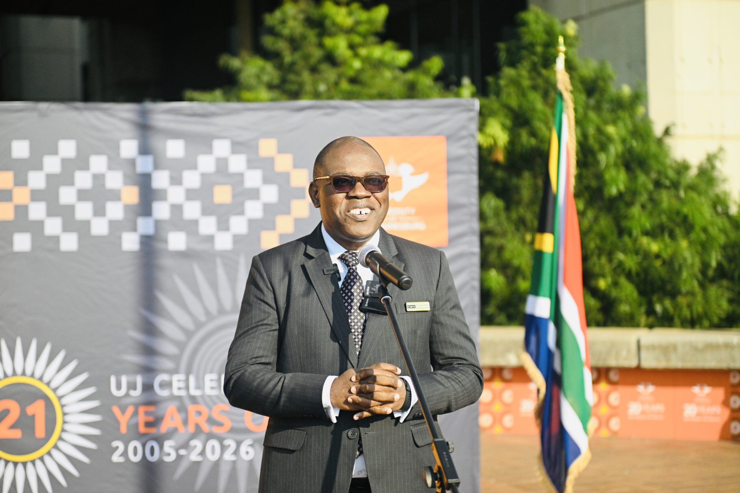 Man in a suit delivering a speech at an outdoor event, with a banner and South African flag behind him.