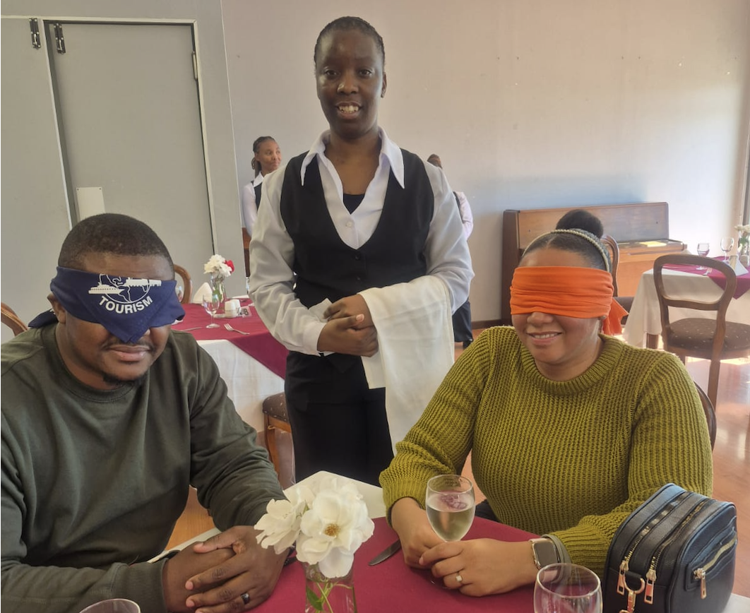 Two diners wear blindfolds at a table as a waiter stands behind them holding a folded napkin in a dining room setting with flowers on the table.