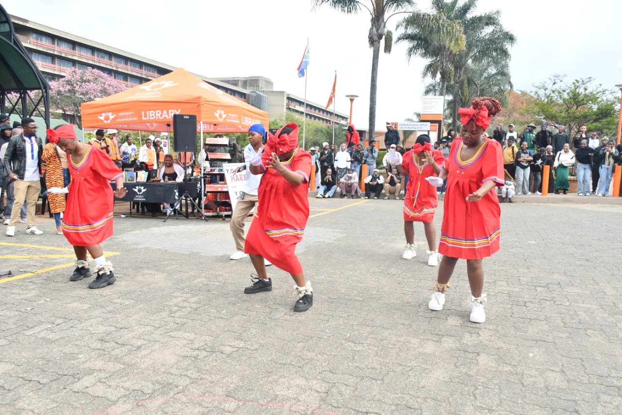 Four women in red dresses dance in a parking lot at a library event, with an orange library tent and a crowd watching in the background.
