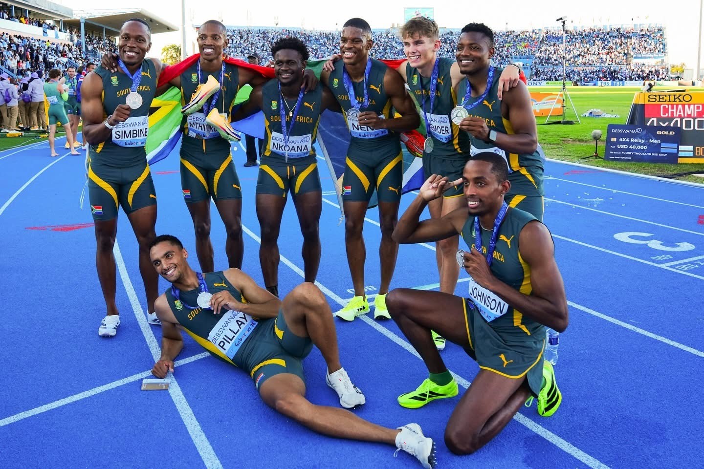 South Africa men's sprint team posing with medals on a blue track, arm-in-arm celebration with two teammates kneeling in front as a stadium crowd looks on.