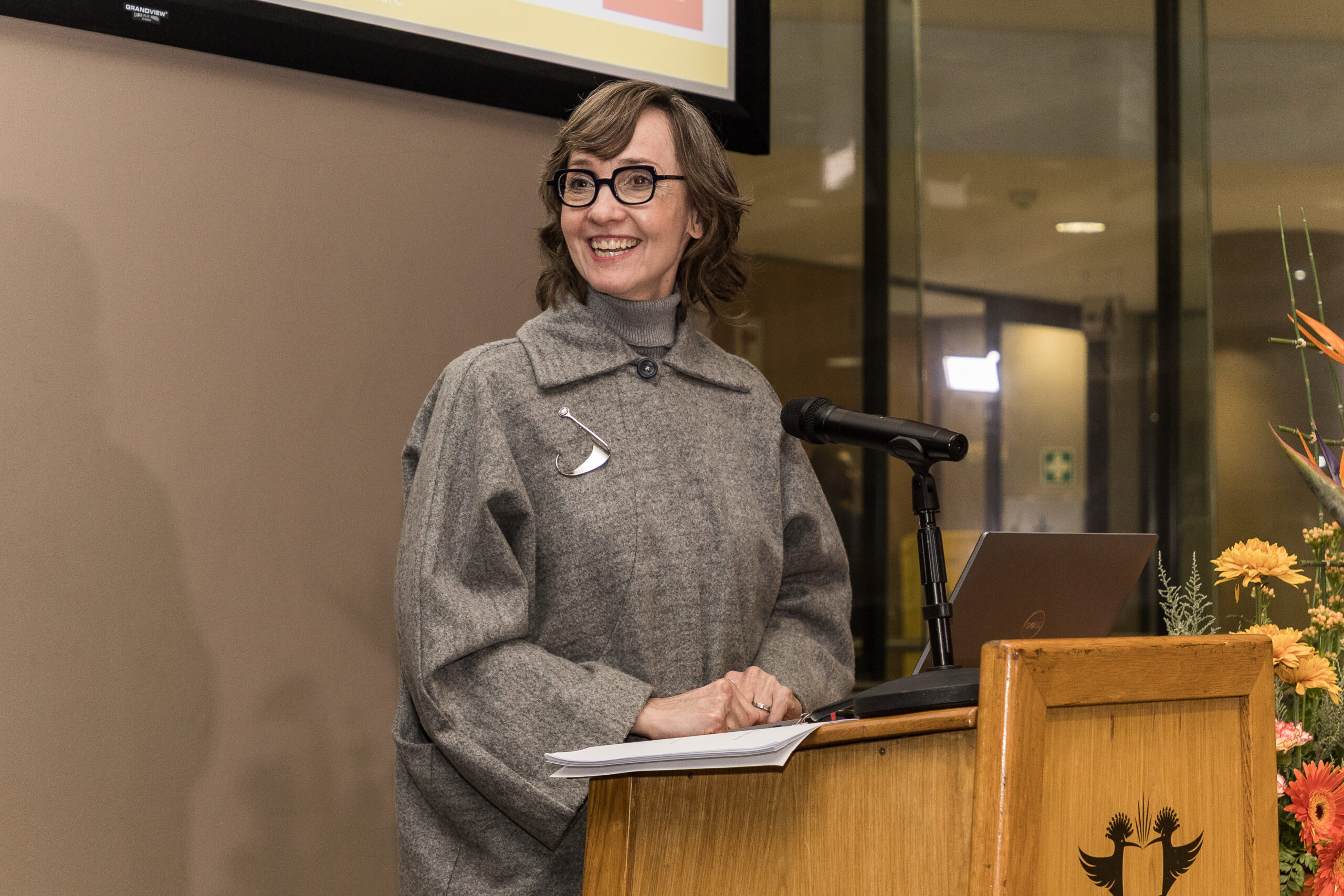 Smiling woman in gray coat speaks at a wooden podium with microphone and papers in front of her.