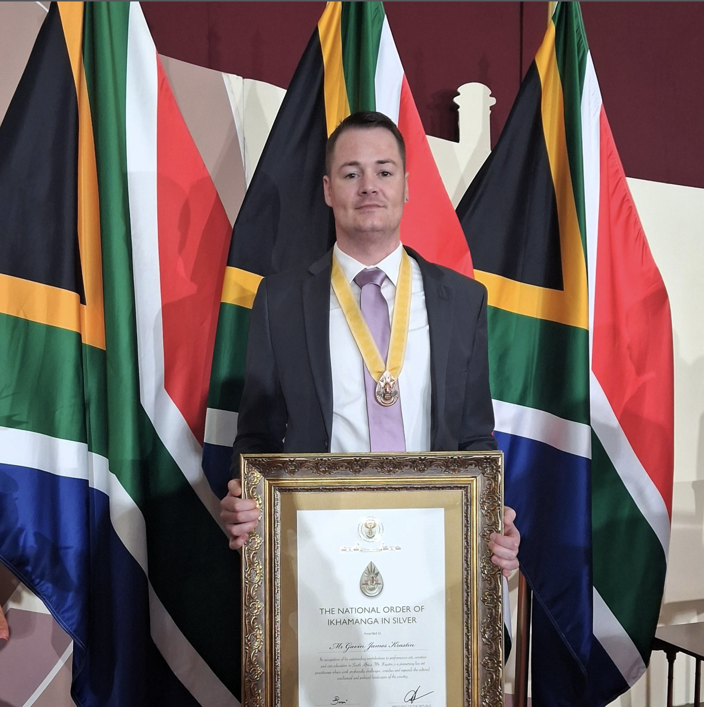 Man in a suit with a gold medal around his neck, holding a framed certificate at an award ceremony with multiple flags in the background.