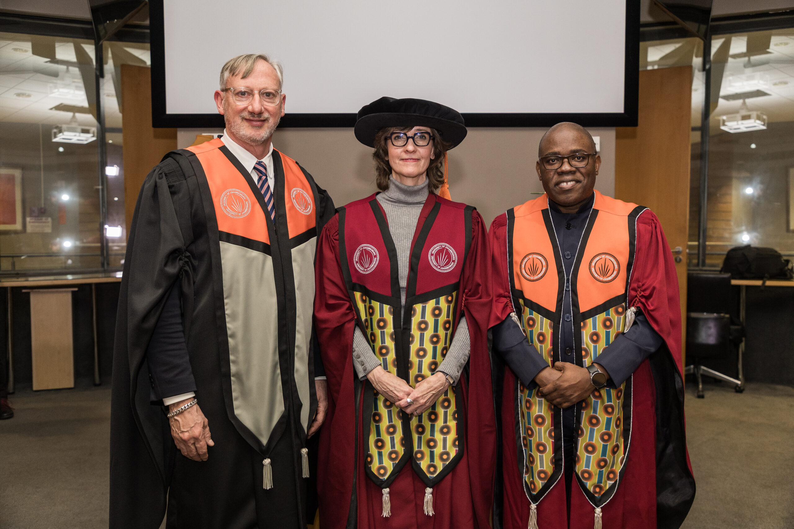 Three graduates in ceremonial robes stand side by side indoors, wearing colorful regalia and caps for a formal ceremony.