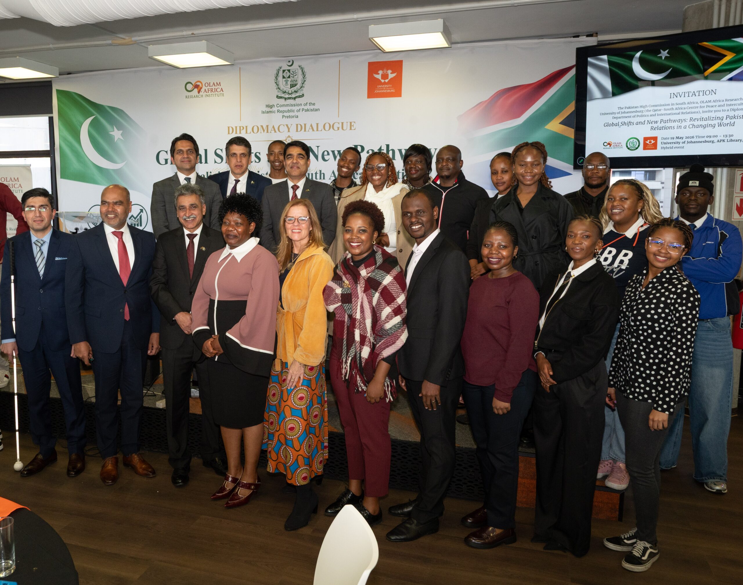 Diverse group posing for a group photo at a diplomacy dialogue event, with banners and flags in the background.