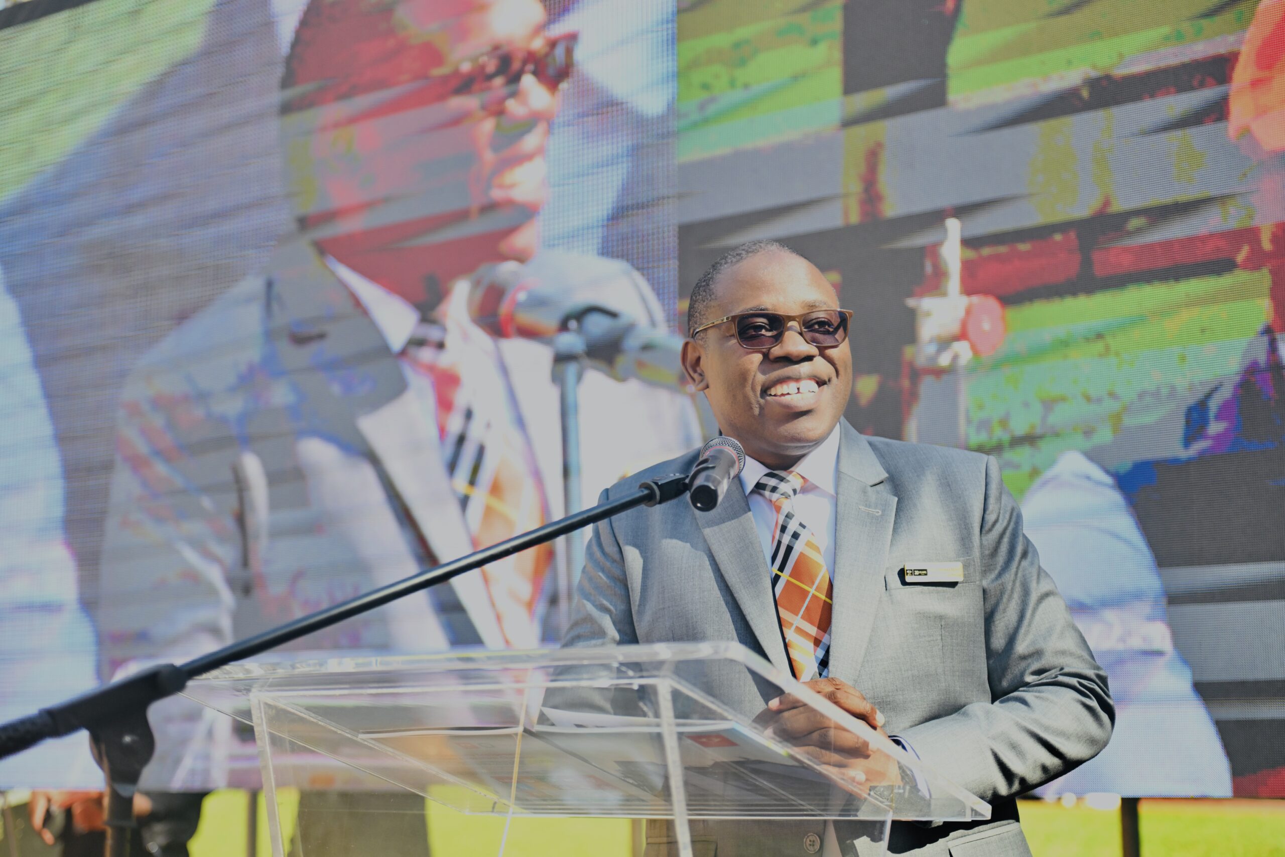 Smiling man in a gray suit and orange striped tie speaks at a clear podium with a microphone against a colorful stage backdrop.