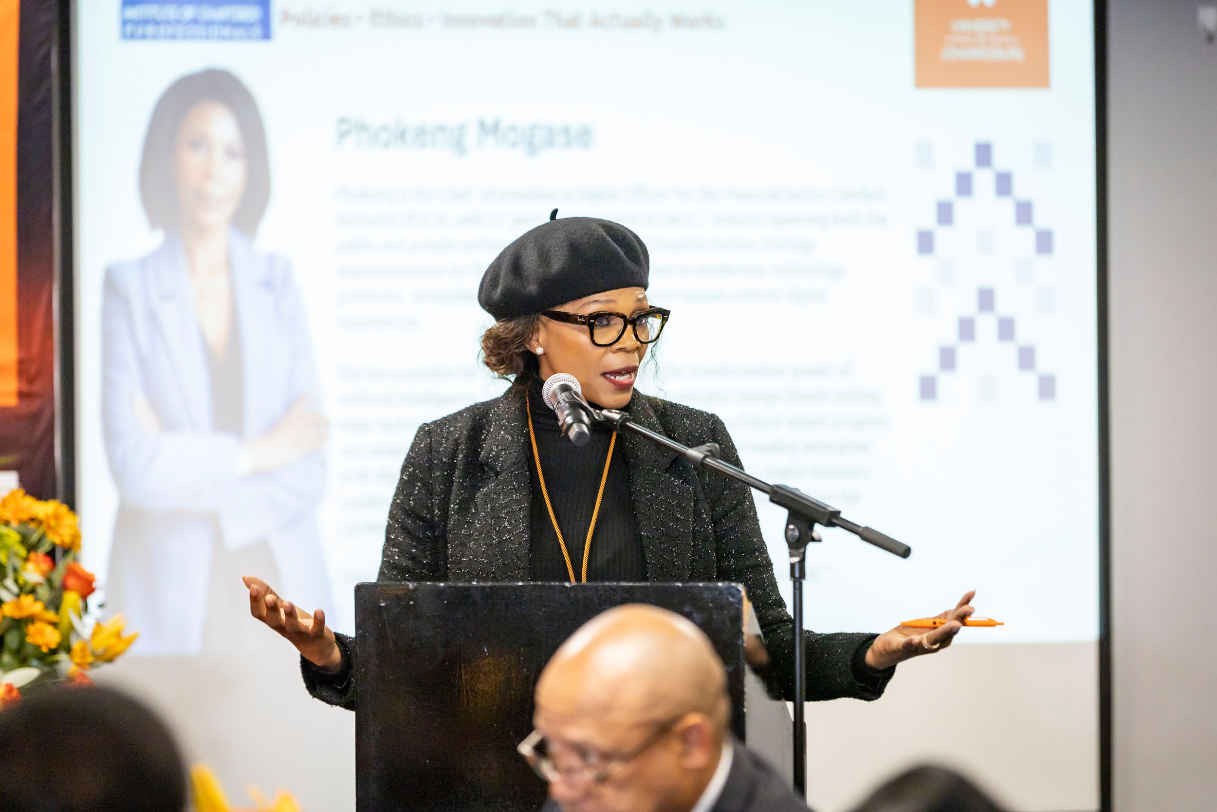 Woman wearing a black beret and glasses speaks at a podium, gesturing with her hands; presentation screen behind her.