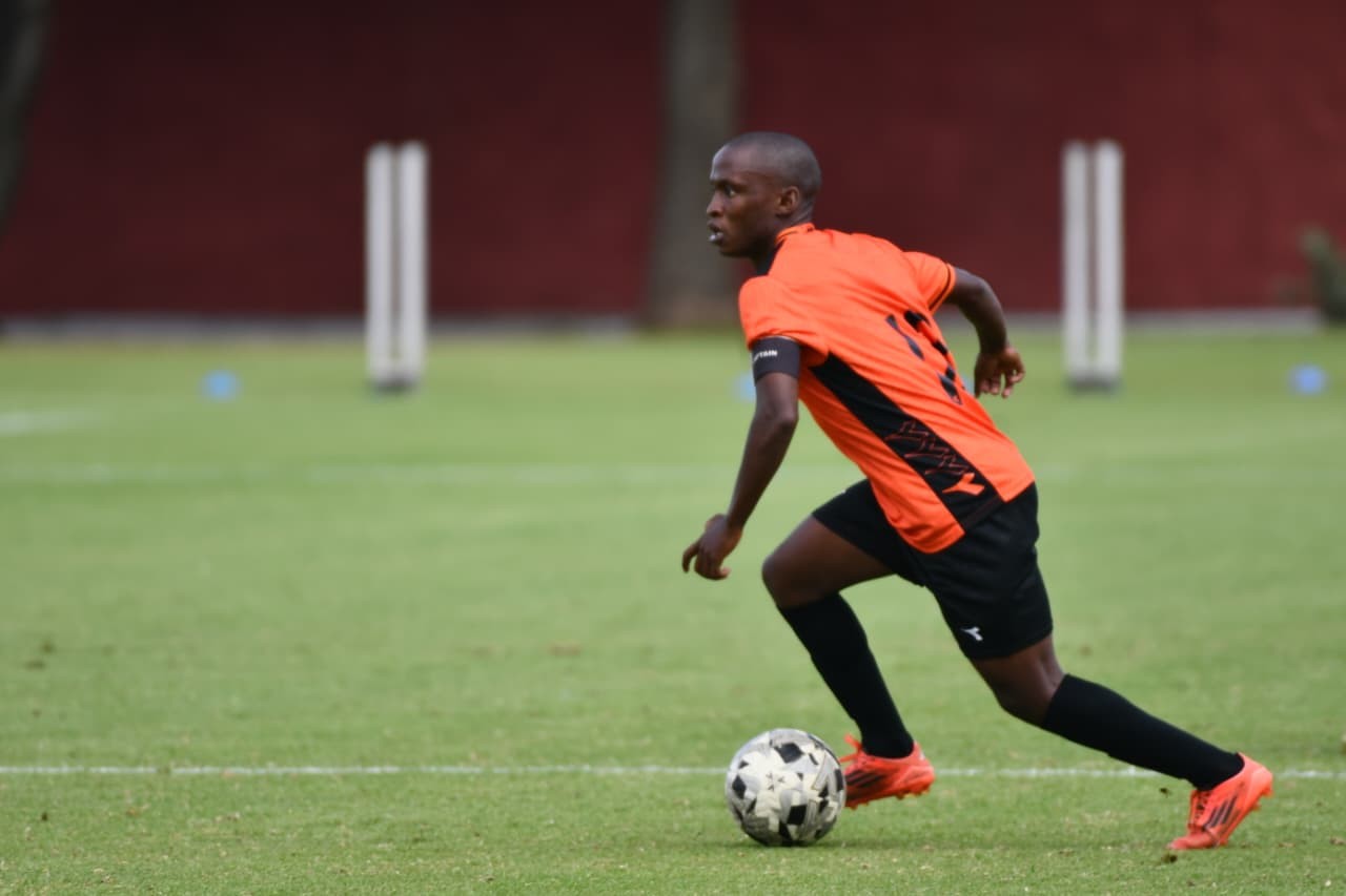 Soccer player in an orange jersey dribbling a ball on a green field, wearing black shorts and bright orange cleats.