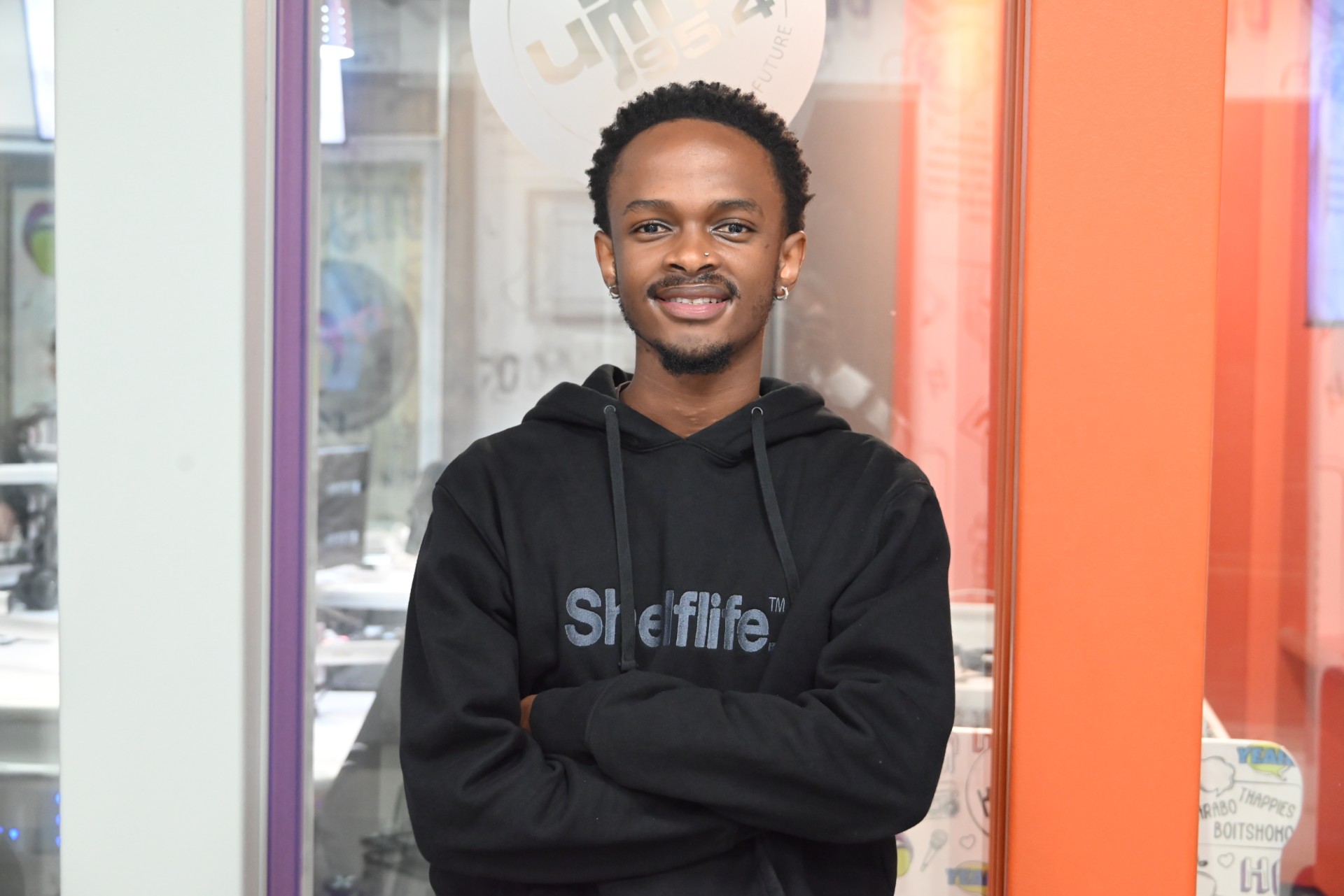 Young man with short curls in a black hoodie with 'ShelfLife' logo, arms crossed, standing in a glass storefront.