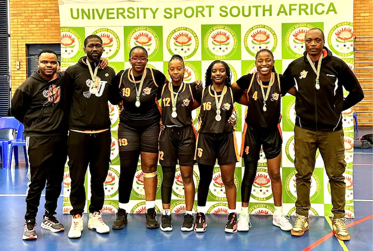 Seven basketball players and coaches pose with medals in front of a University Sport South Africa banner.”